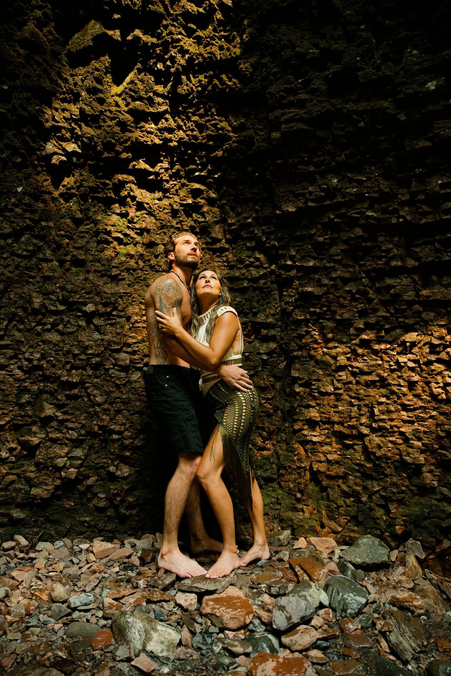 Couple embracing barefoot against ancient stone wall in dramatic cave-like setting with warm lighting.
