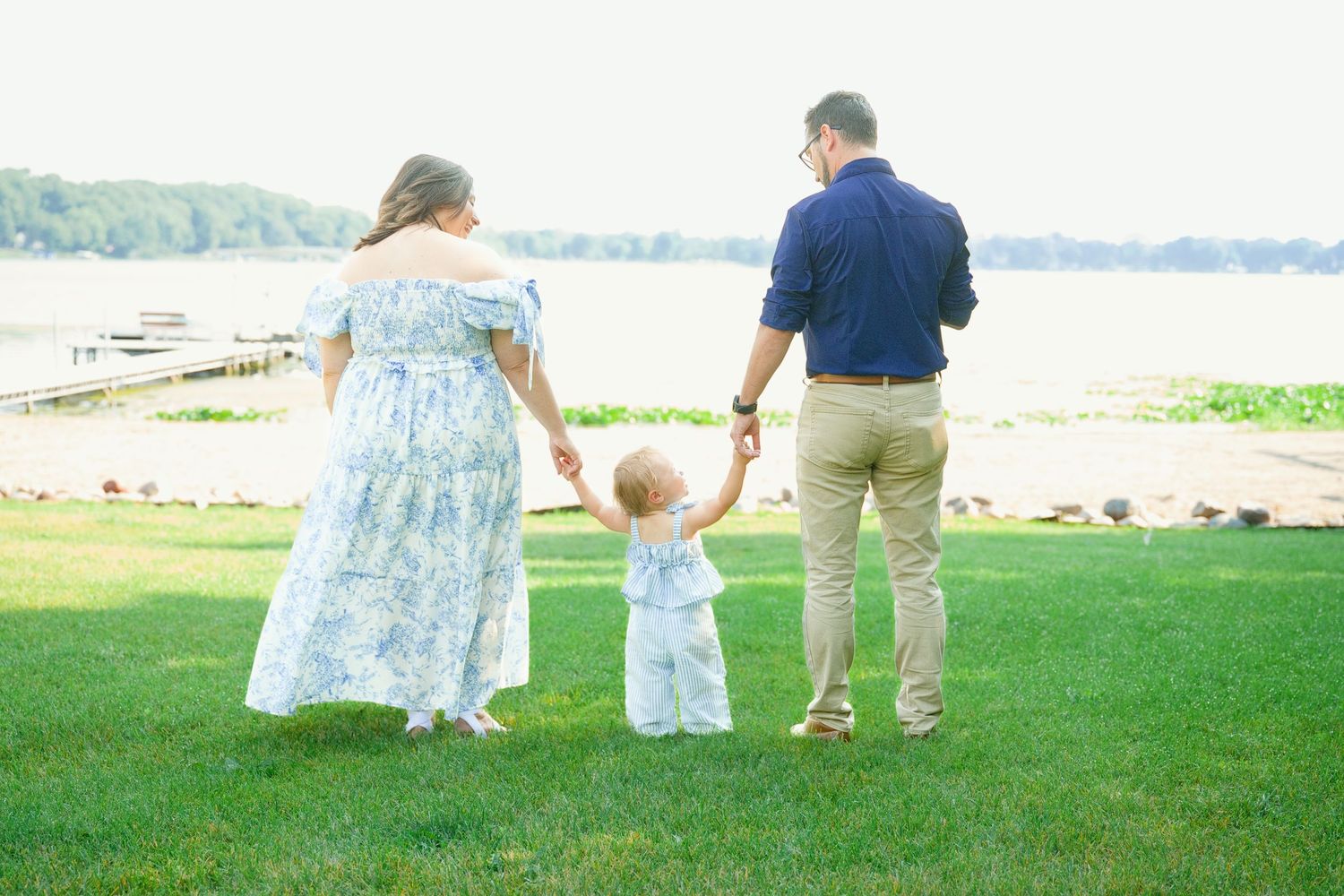 Young family holding toddler's hands while walking on grass near waterfront on sunny day.