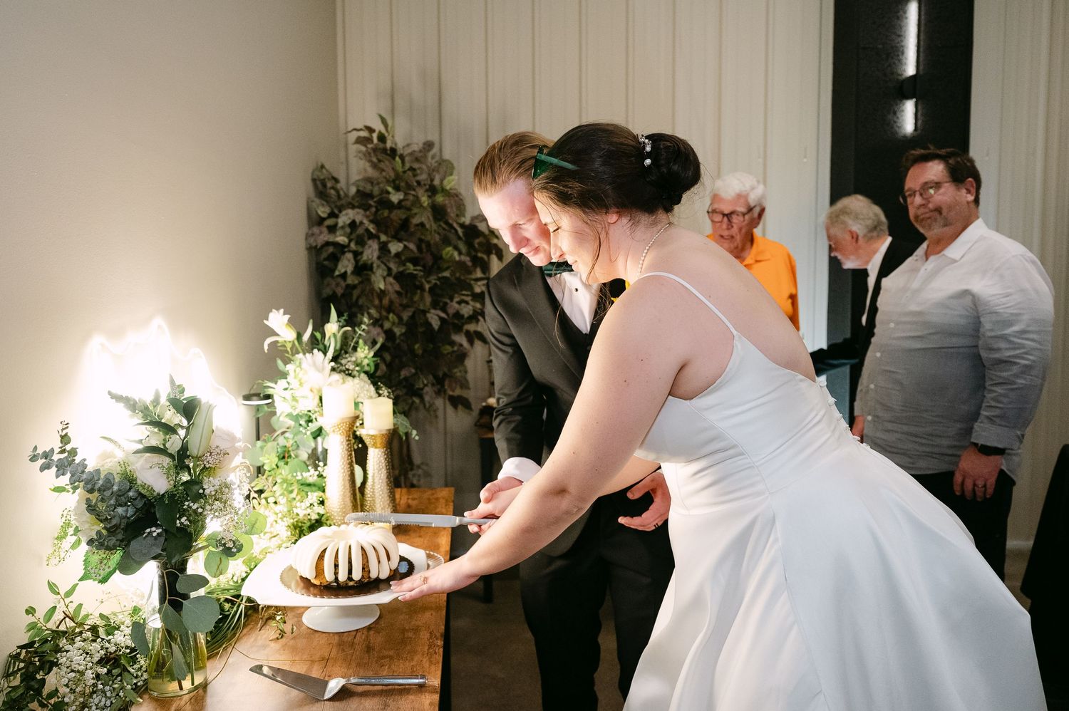 A couple in wedding attire cuts their bundt cake together at an intimate indoor reception with greenery decor.