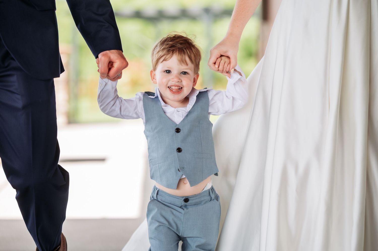 A ring bearer in a light blue vest walks beside a bride and groom during an outdoor wedding ceremony. Pease Minnesota Wedding at Northern Li