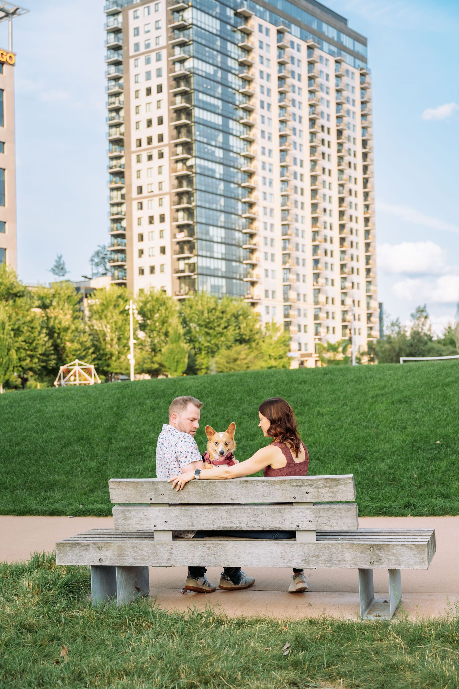 A couple sits together on a wooden bench against a backdrop of modern high-rise apartment buildings.