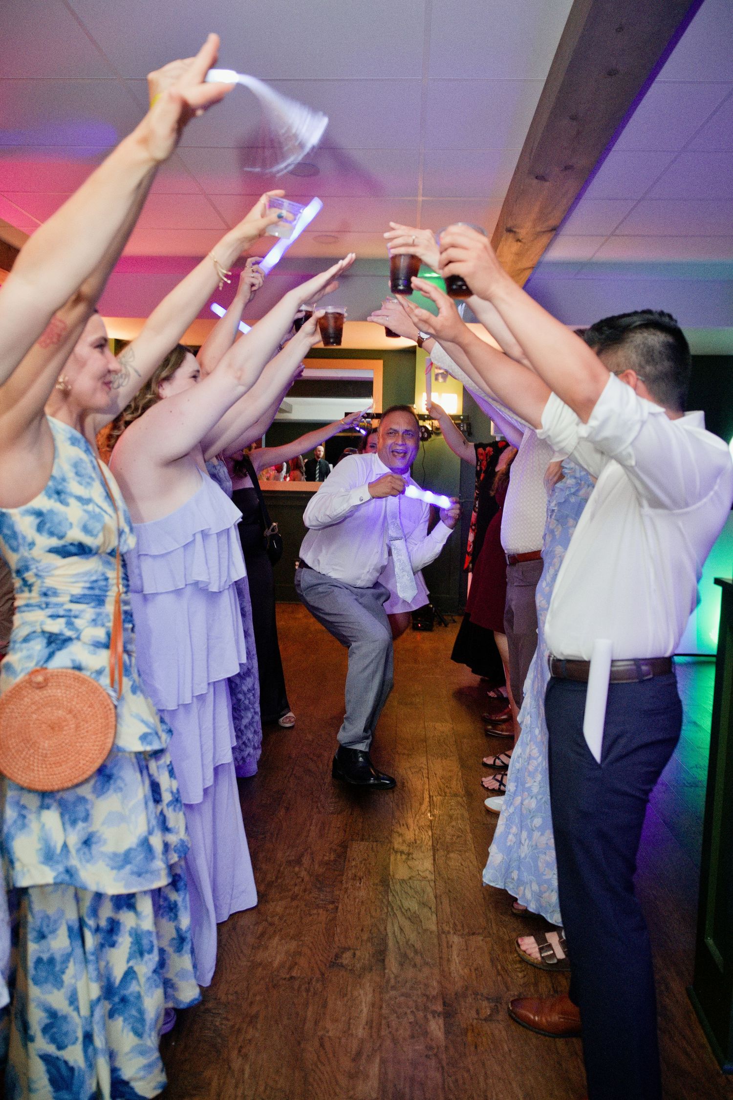 Wedding guests raise glasses in a celebratory tunnel as a man dances through at a festive indoor reception.
