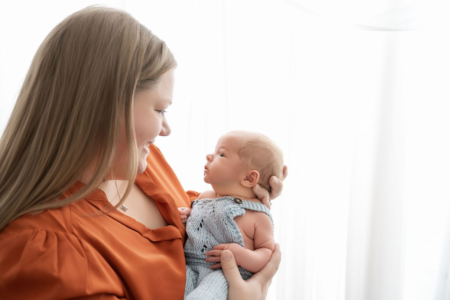 Minnesota Best newborn photo a Parent in orange sweater tenderly holds and nurtures small baby in soft grey onesie against white background.
