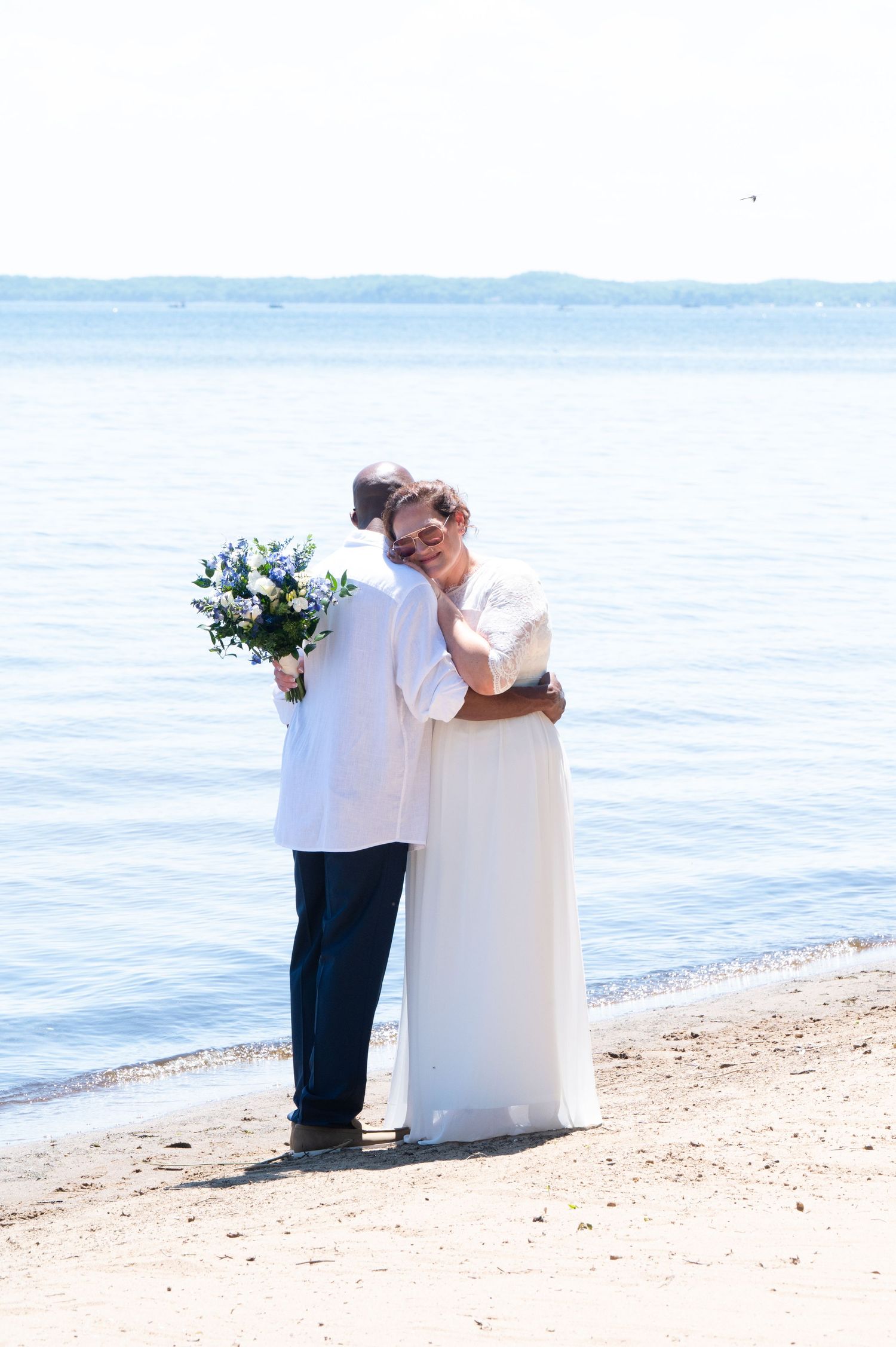 Bride and groom embracing on a beach shoreline holding a blue and white bouquet.
