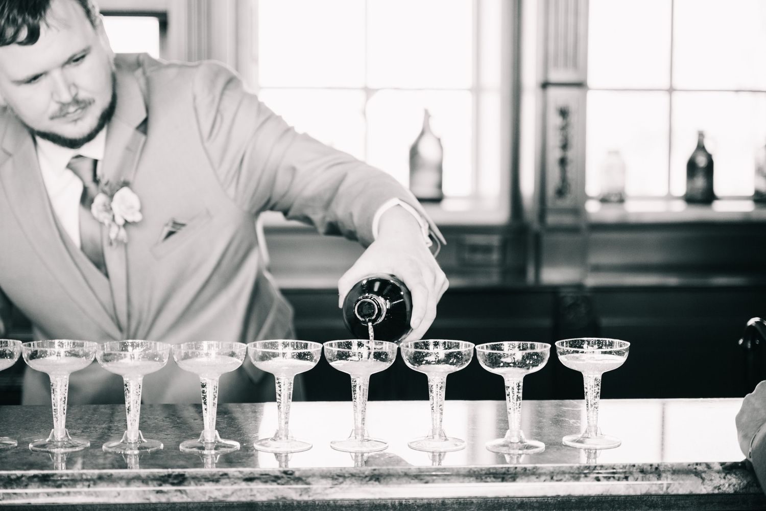 Black and white photo of a groom pouring champagne into a row of coupe glasses at a bar.