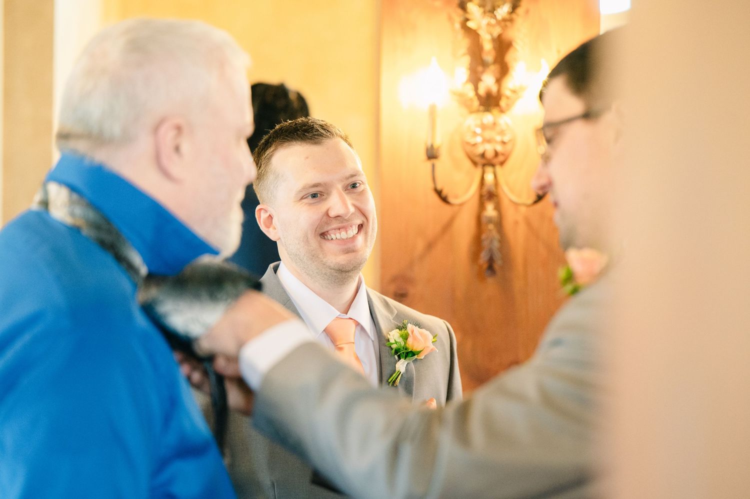 Smiling groom in gray suit having boutonniere pinned while chatting with guests before wedding ceremony.