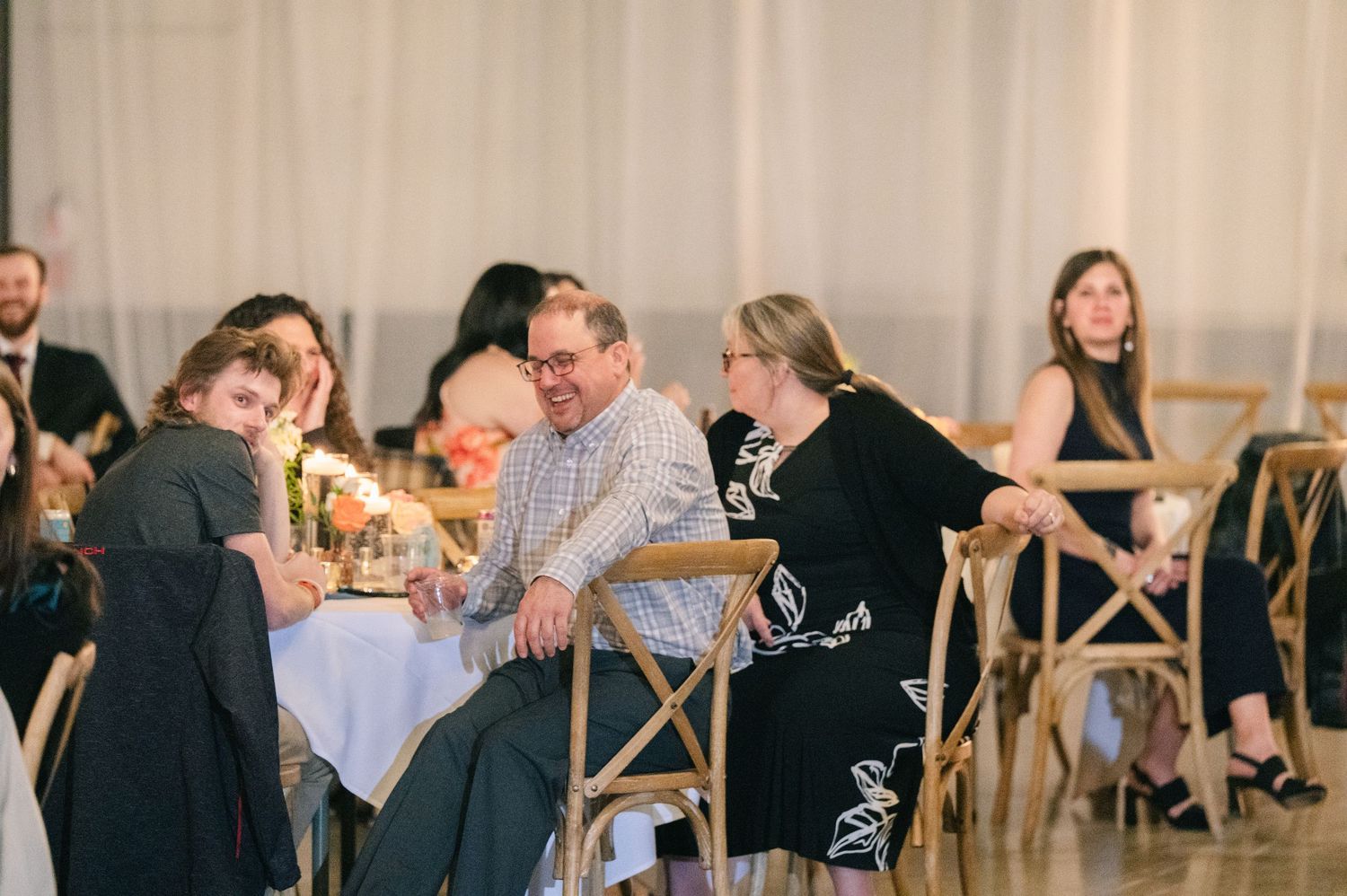 Wedding guests laugh and chat together at candlelit reception table in draped venue space.