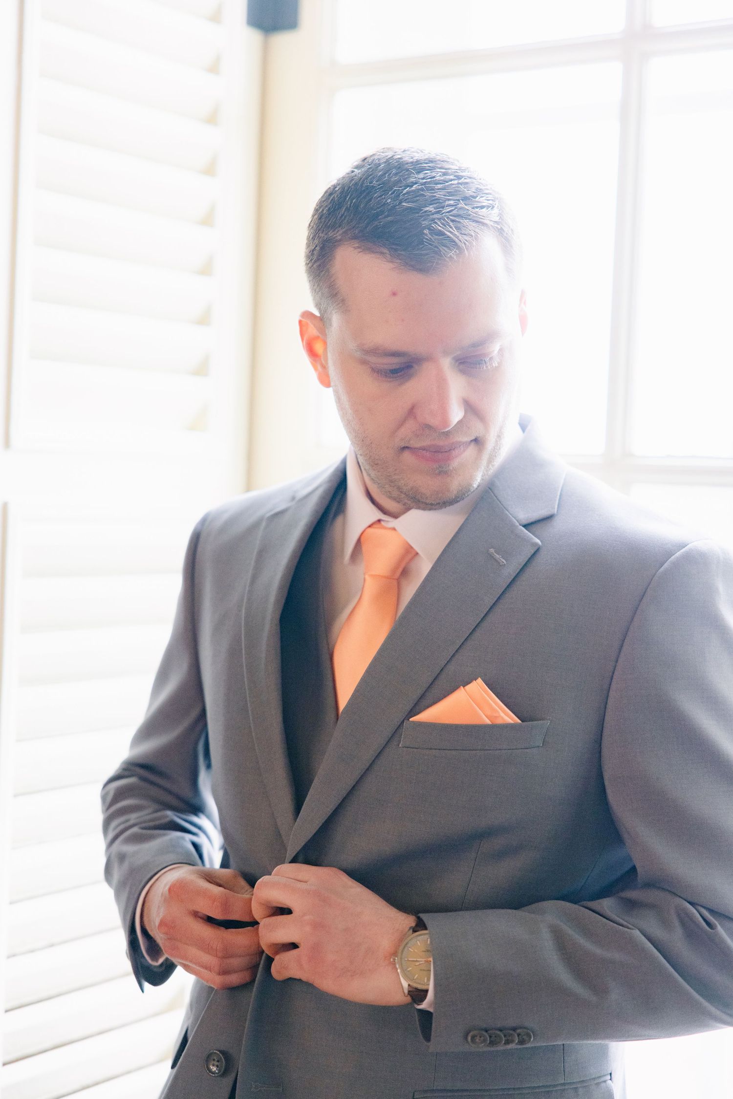 Groom in gray suit with peach tie and pocket square buttoning jacket near bright window.
