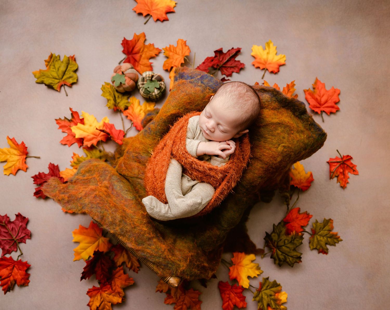 Artistic autumn photography setup with warm brown blankets and orange maple leaf decorations against a neutral backdrop.