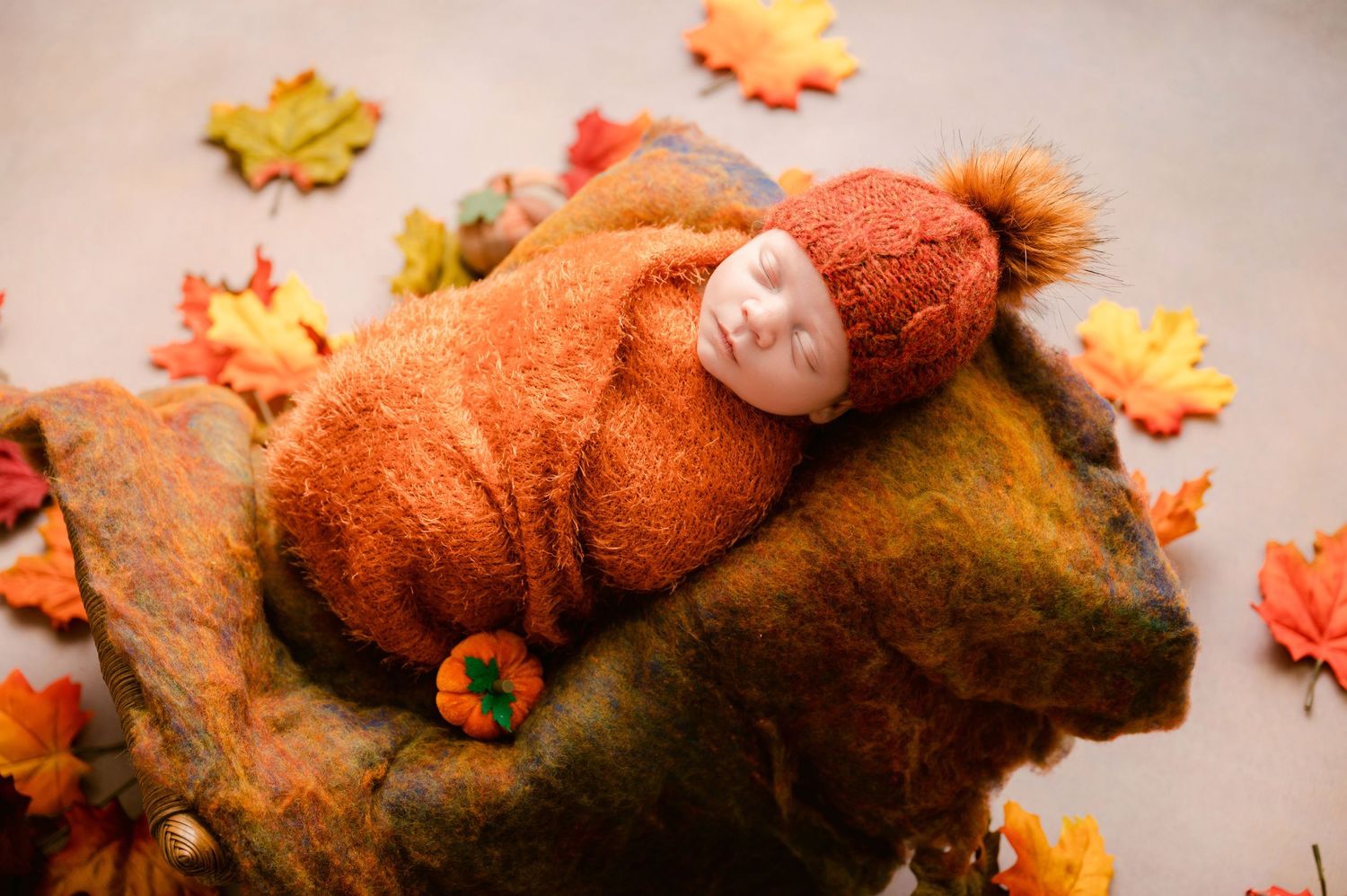 A sleeping baby dressed in an orange knit outfit rests on a bed surrounded by autumn maple leaves.