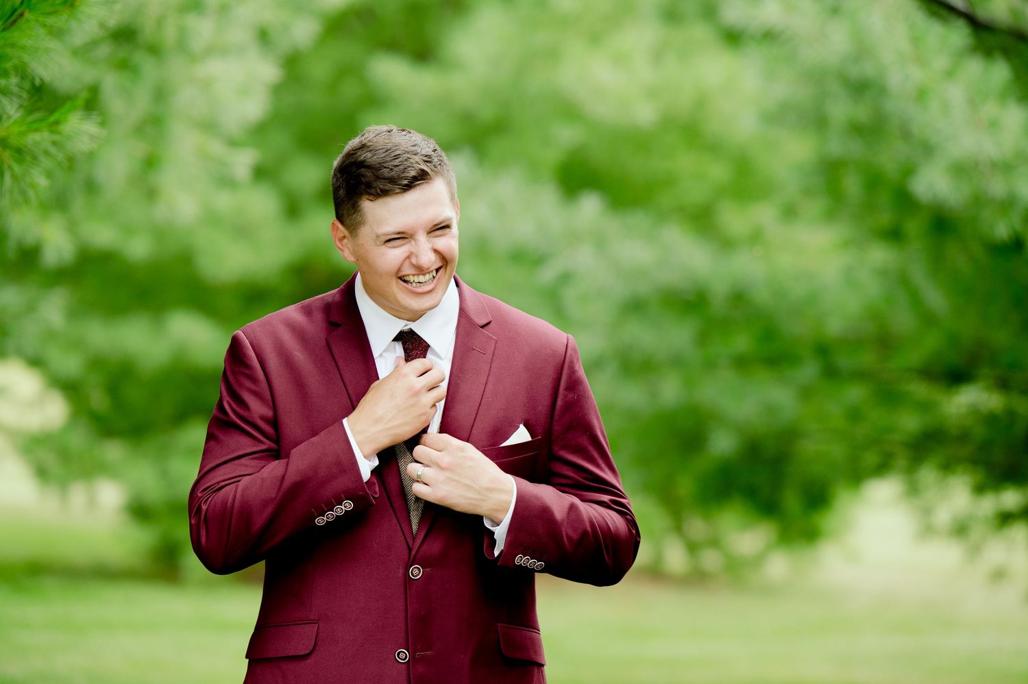 A groom in a burgundy suit adjusts his tie while smiling outdoors.