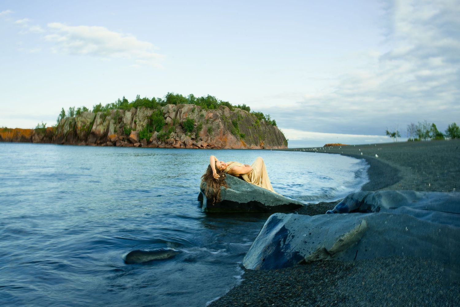 Driftwood on a rocky black sand beach with a forested island in calm blue waters under a partly cloudy sky.