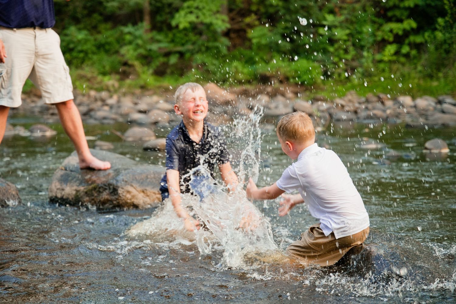 Two children joyfully splashing water in a rocky stream outdoors on a sunny day.