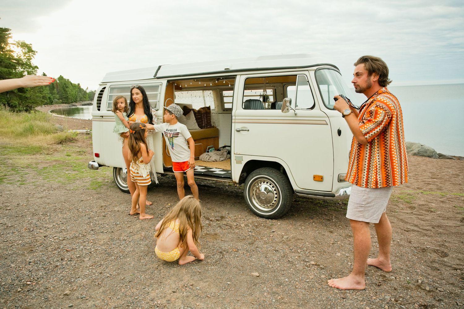 Family posing by a vintage VW camper van on a rocky shore, man photographing woman and children near open sliding door.
