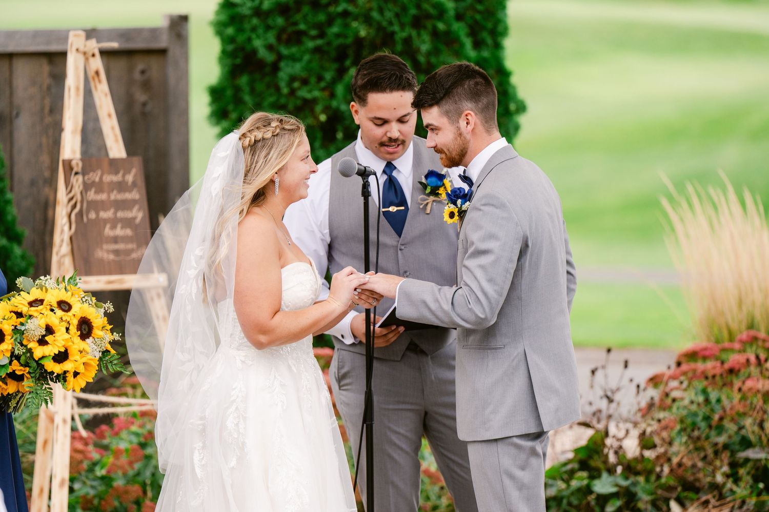 Central MN Wedding Photos - Bride and groom exchange rings outdoors with officiant, sunflowers and rustic wooden sign in background.