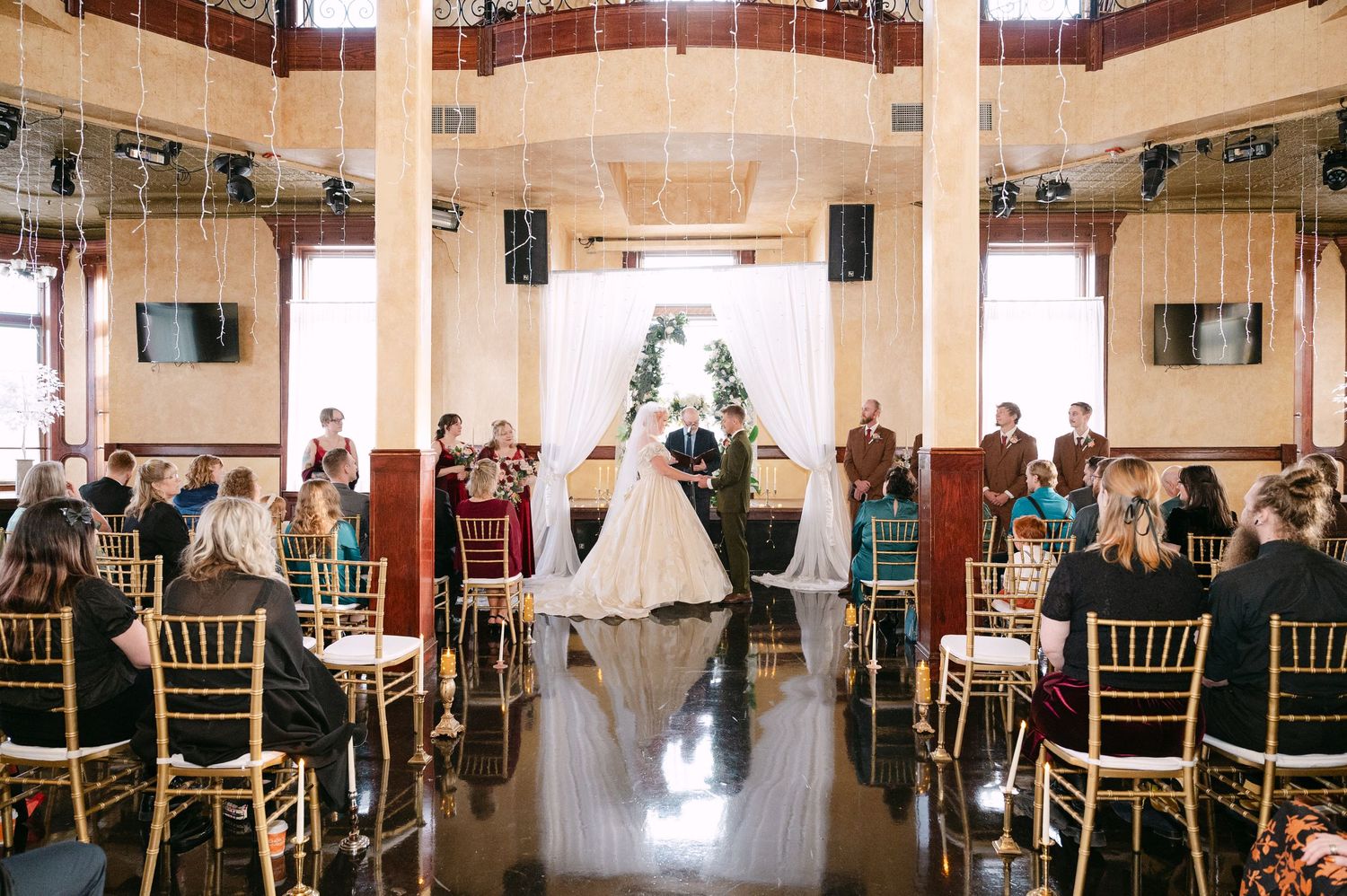Central MN Wedding Photos | Wedding ceremony with couple at the altar in an elegant ballroom with gold chairs and guests seated.