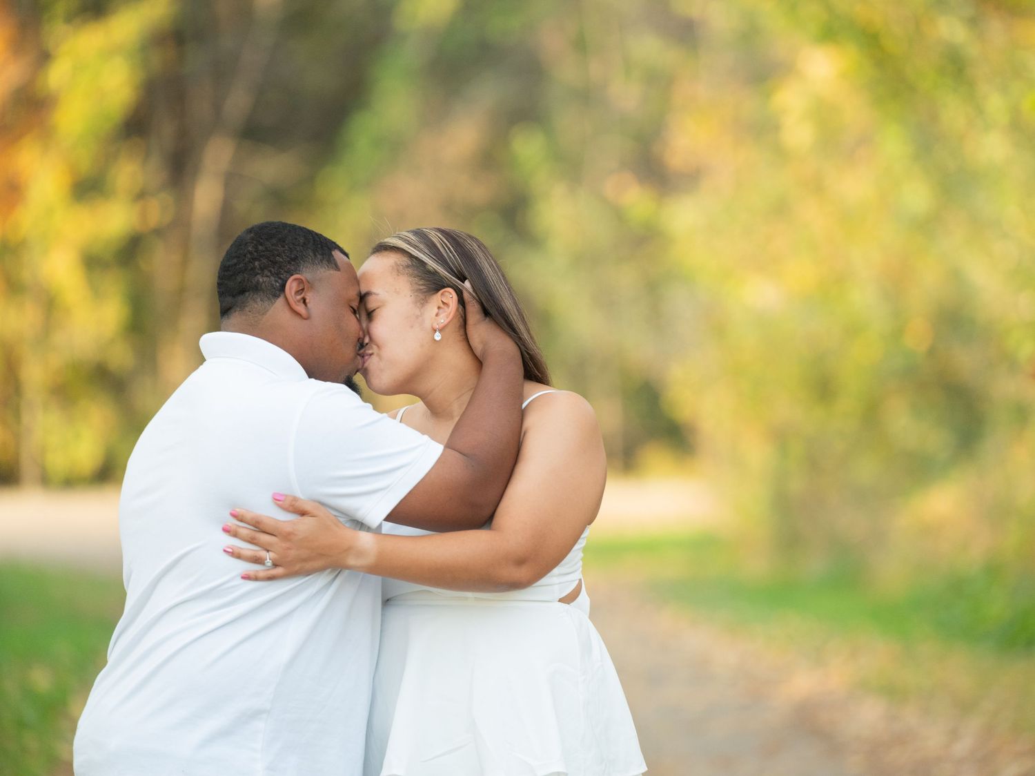Central Minnesota Best Engagement Photographer Couple sharing a romantic kiss outdoors on a tree-lined path in autumn, wearing white outfits