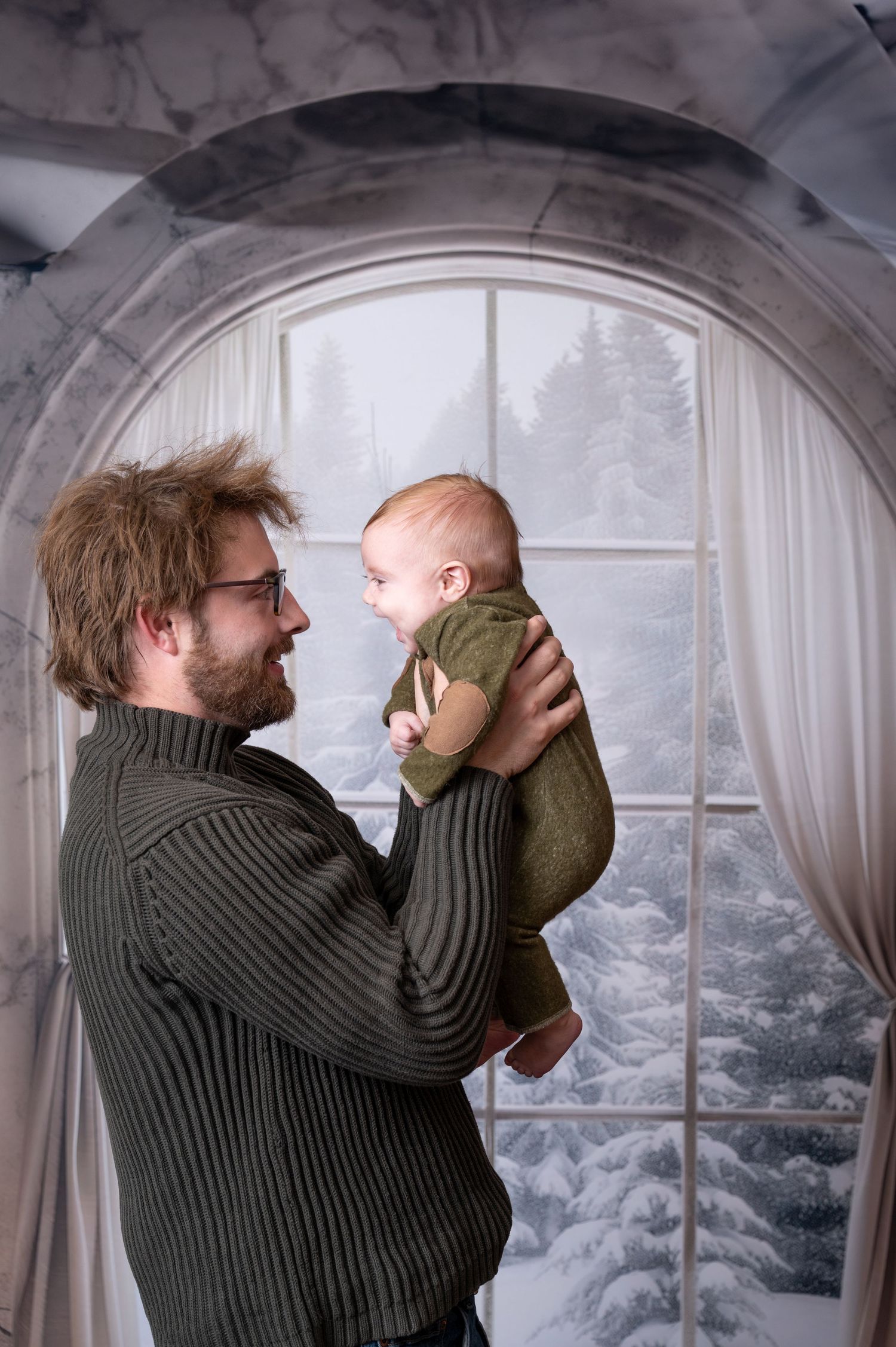 Man in ribbed sweater holds baby by arched window with snowy winter scene visible outside. Princeton MN Milestone, Maternity and Newborn P