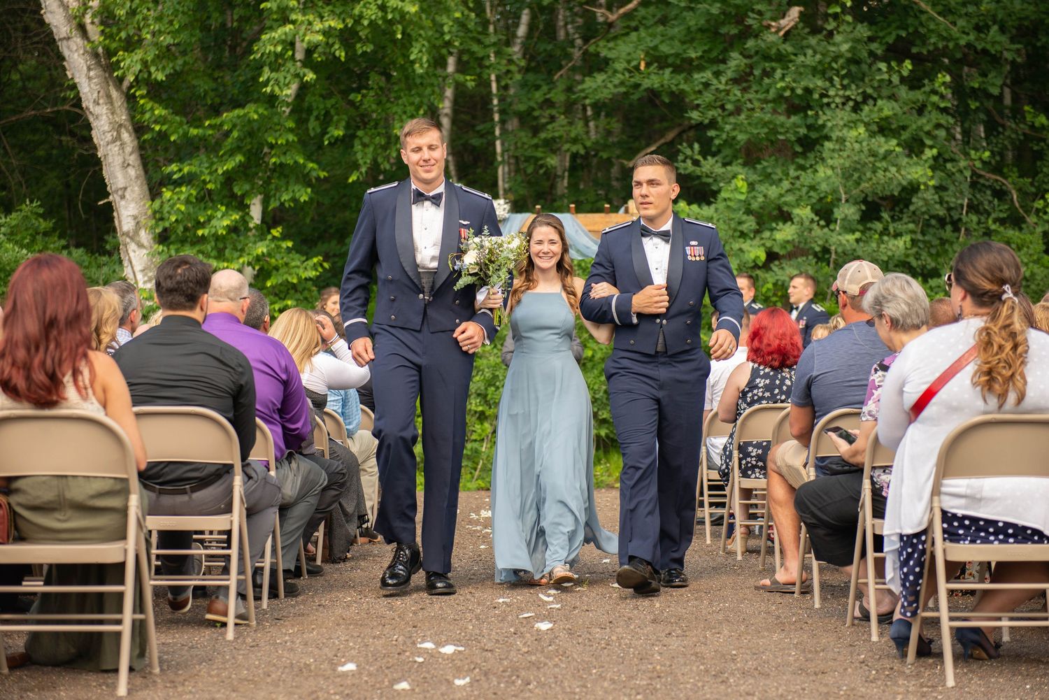 Wedding guests sit on folding chairs at an outdoor ceremony under green trees. Zimmerman MN Weddings