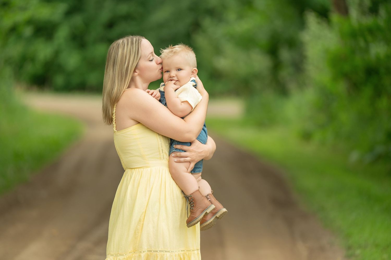 A woman in a yellow sundress holds a baby while standing on a dirt path surrounded by lush greenery.