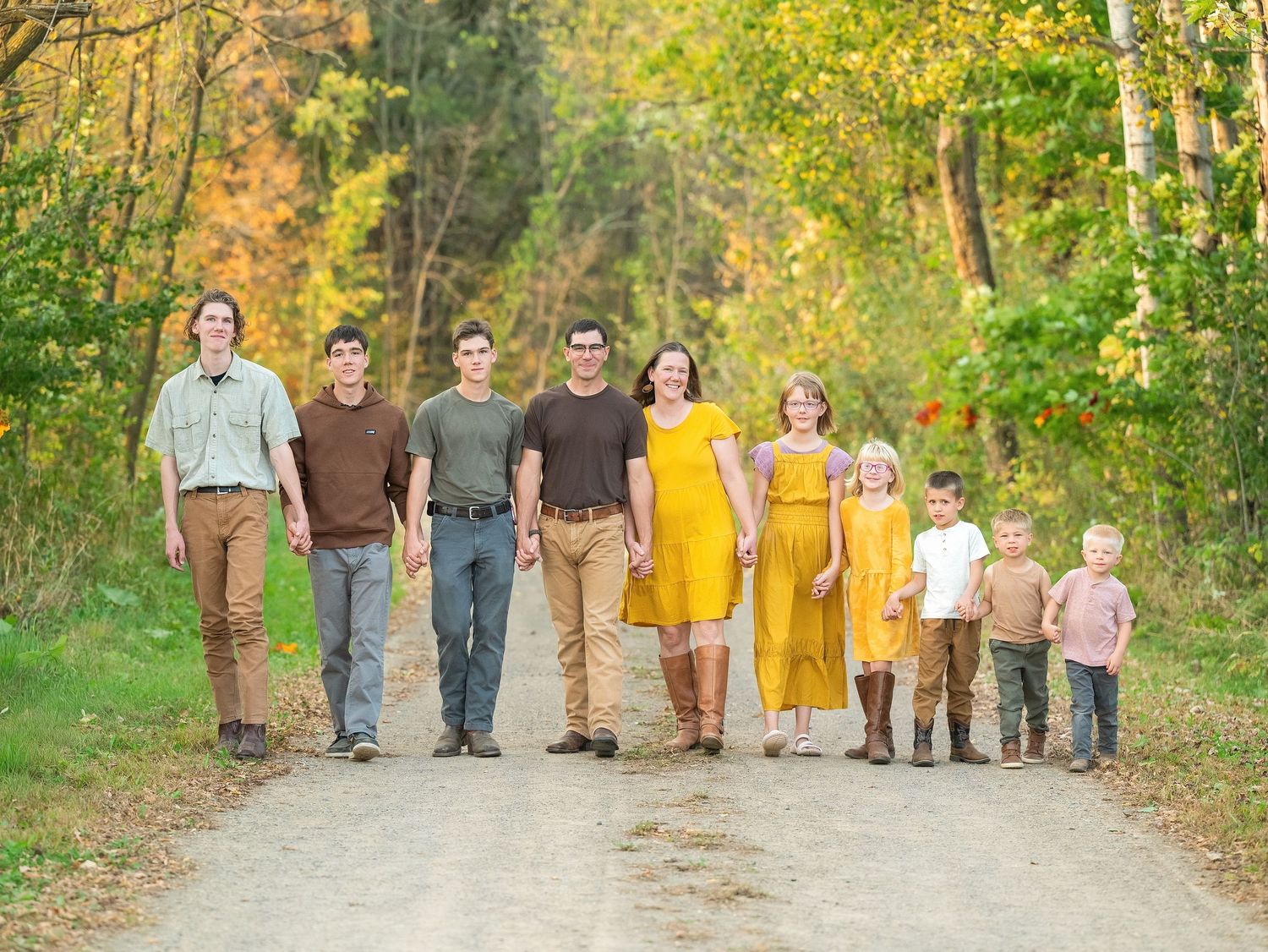 A group of people wearing coordinated earth-toned and yellow outfits stand together on a leaf-covered autumn path.