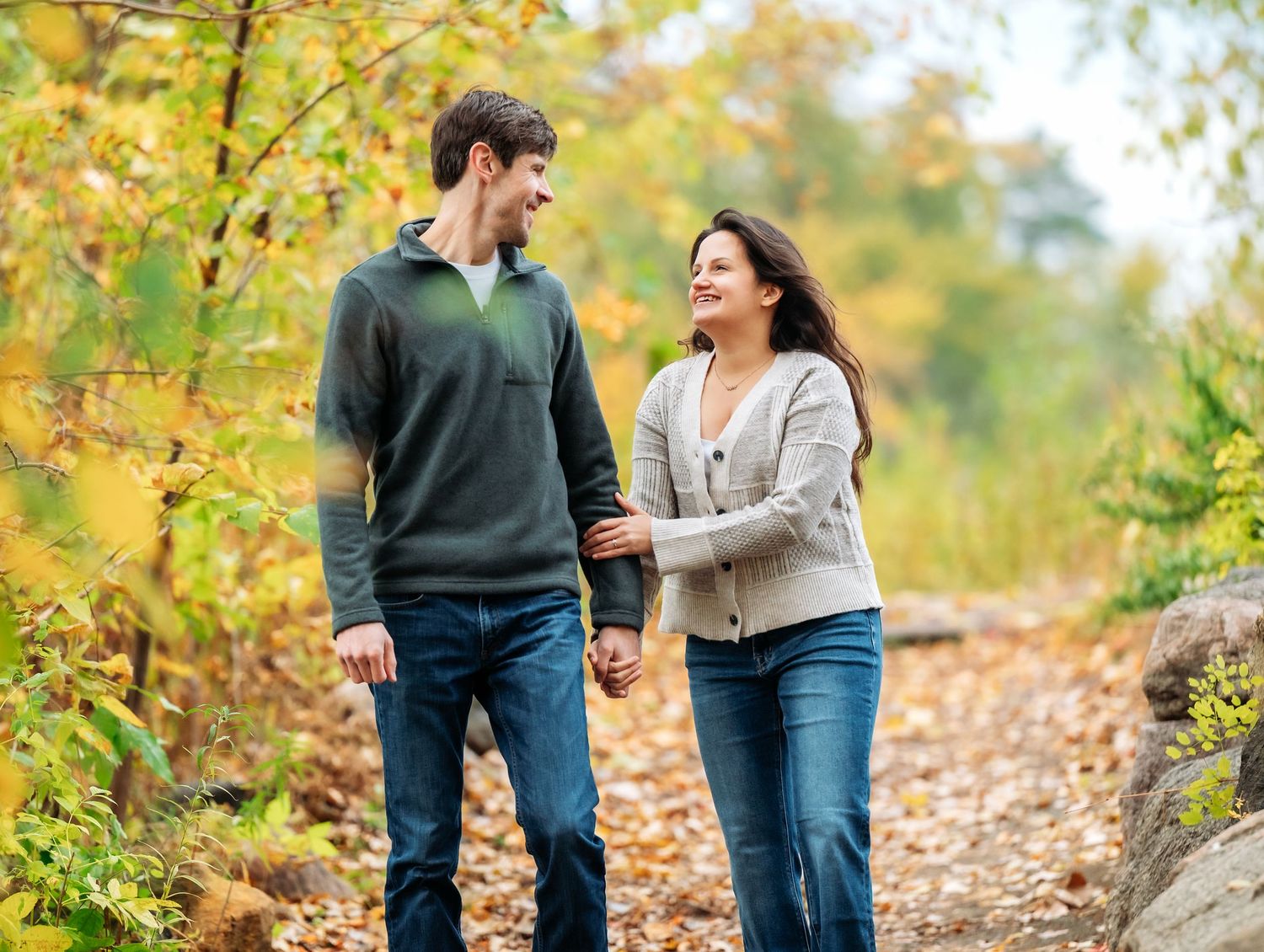 Two people walk together on a leaf-covered path surrounded by fall foliage and trees at Lake Como. Engagement Sessions Minnesota.