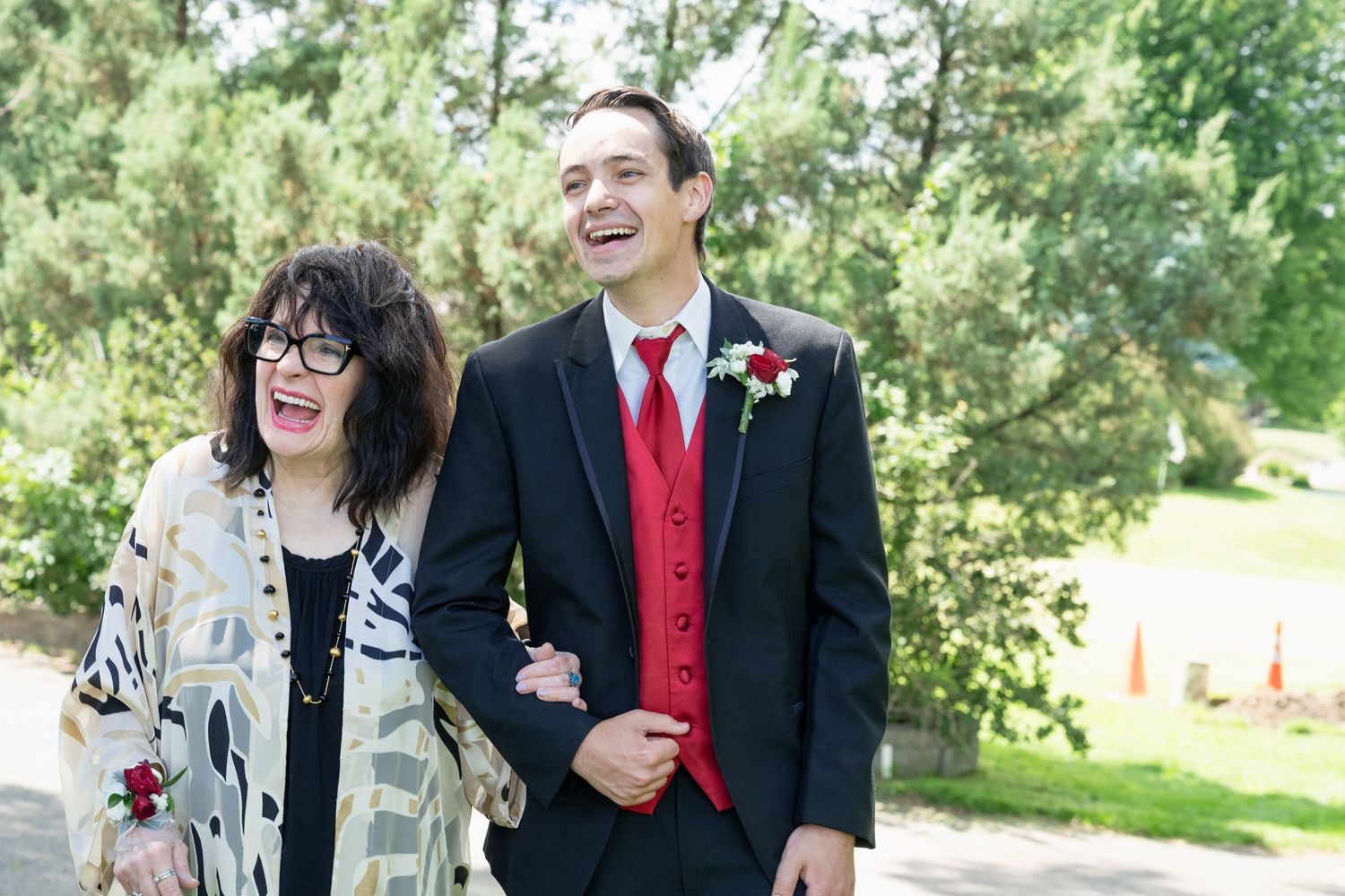 Laughing groom in black tuxedo with red vest walks with smiling woman outdoors.