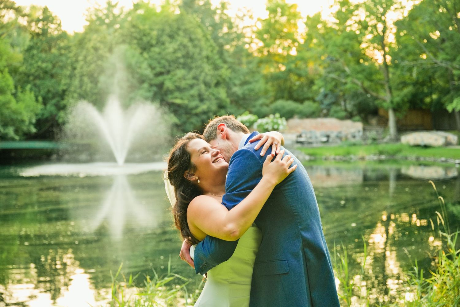 Couple embracing joyfully by a serene pond with a fountain and lush green trees in the background.