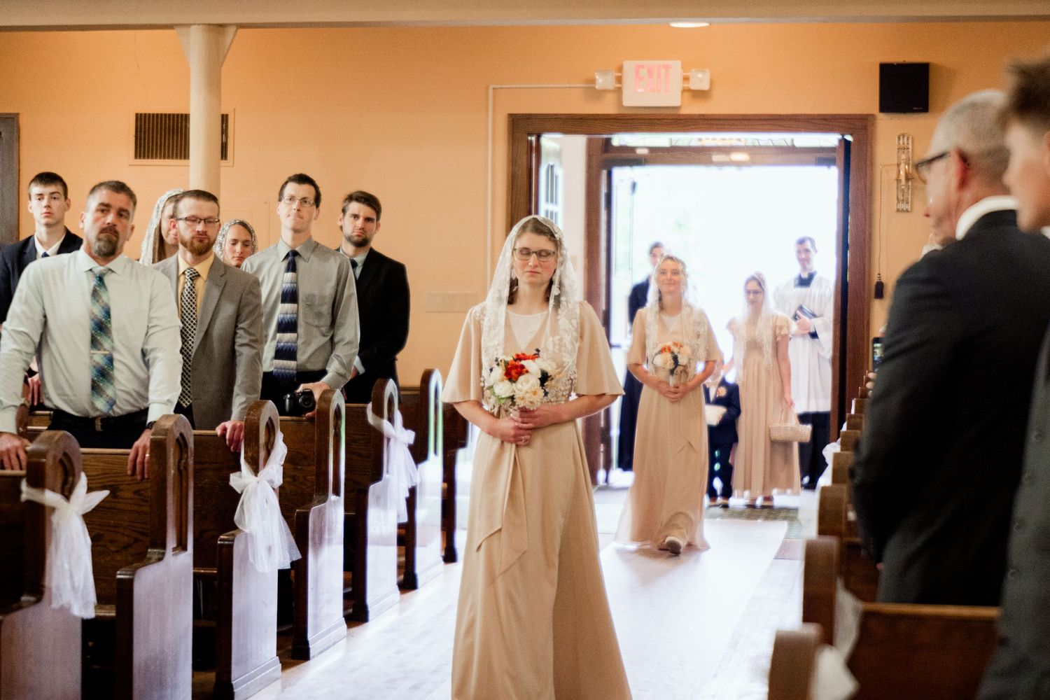 A wedding procession in a church aisle with a bride in a cream gown walking down the pew-lined corridor.