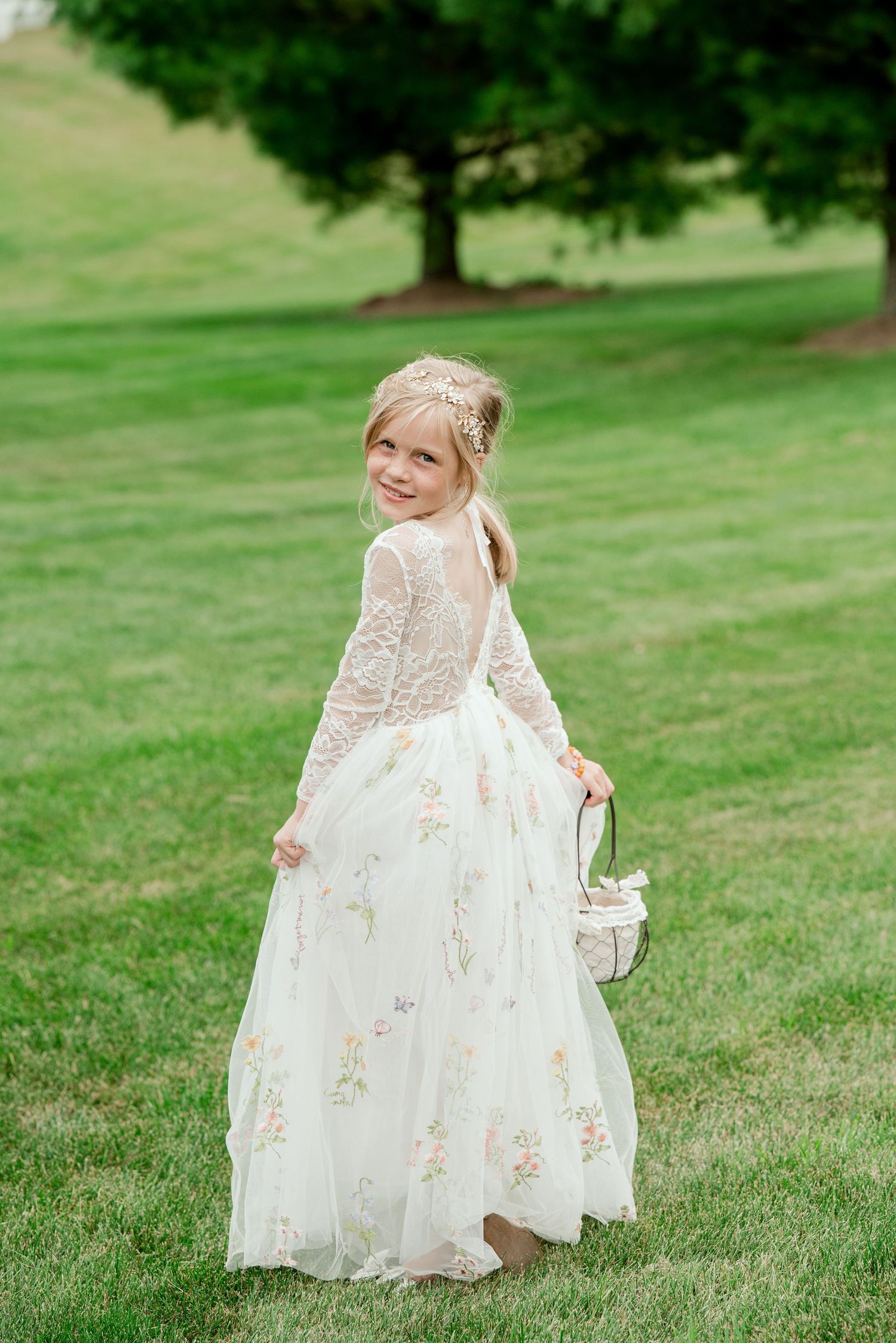 A young flower girl in a delicate white lace dress with floral details stands on a grassy lawn near a tree.