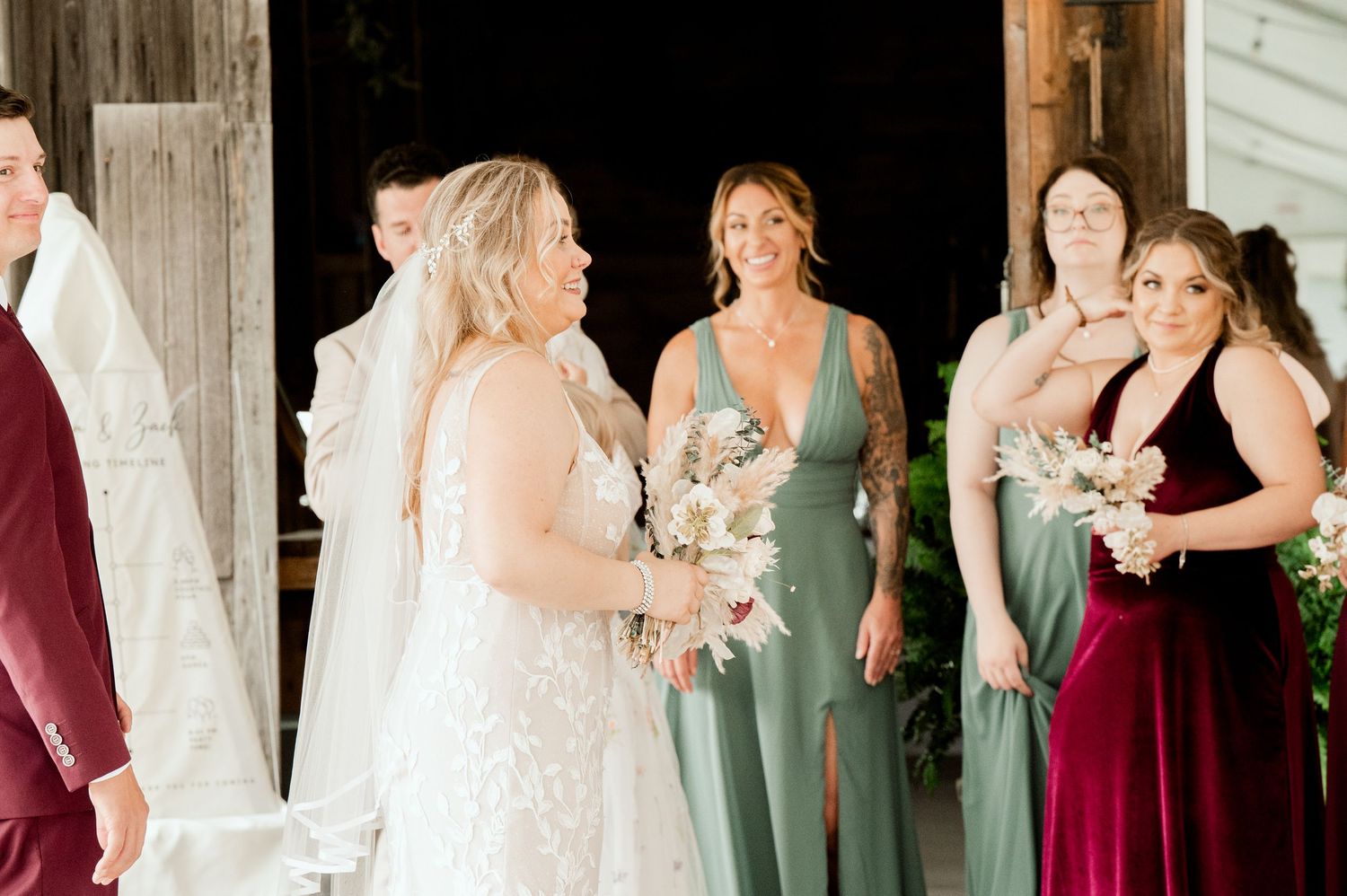 Wedding party gathered in rustic barn doorway wearing burgundy and sage green dresses.