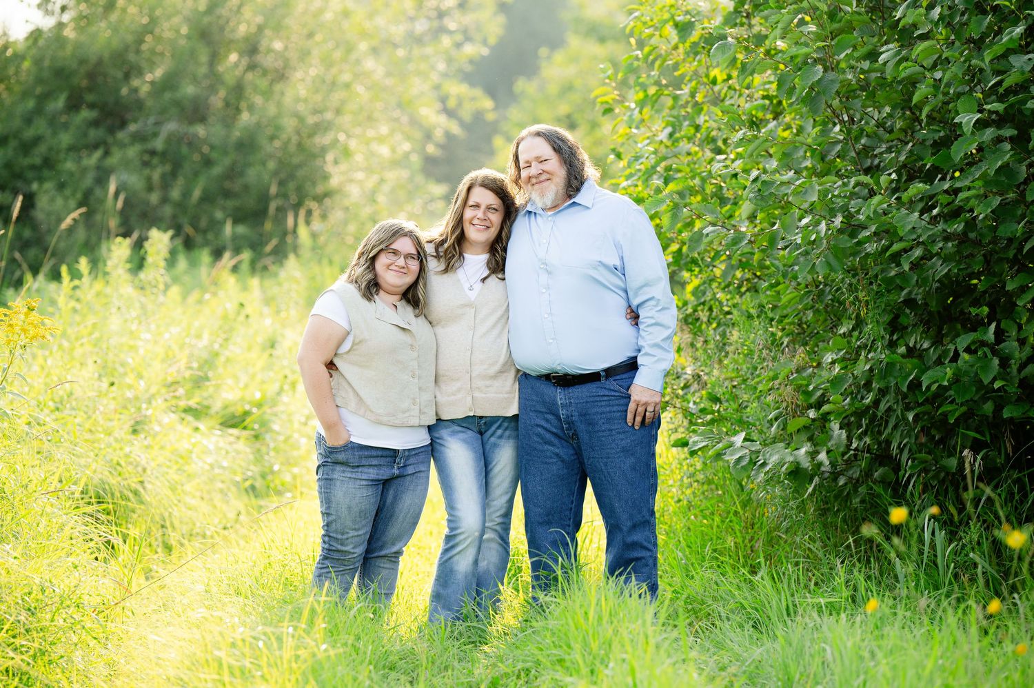 Three people walk together through a lush green field with bright yellow wildflowers on a sunny summer day.
