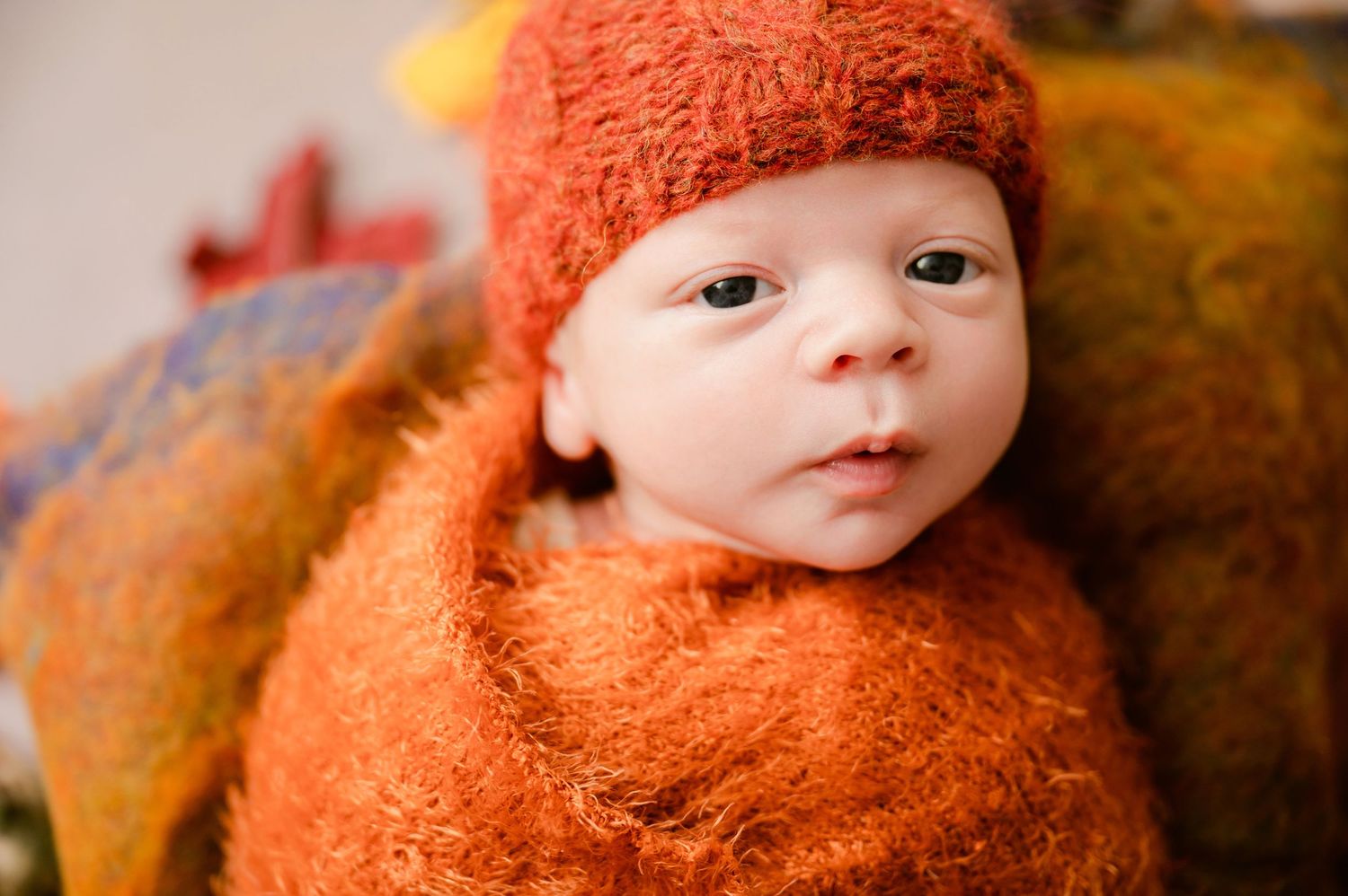 A baby bundled in a cozy orange knit outfit and matching cap lies against an autumn-colored background.