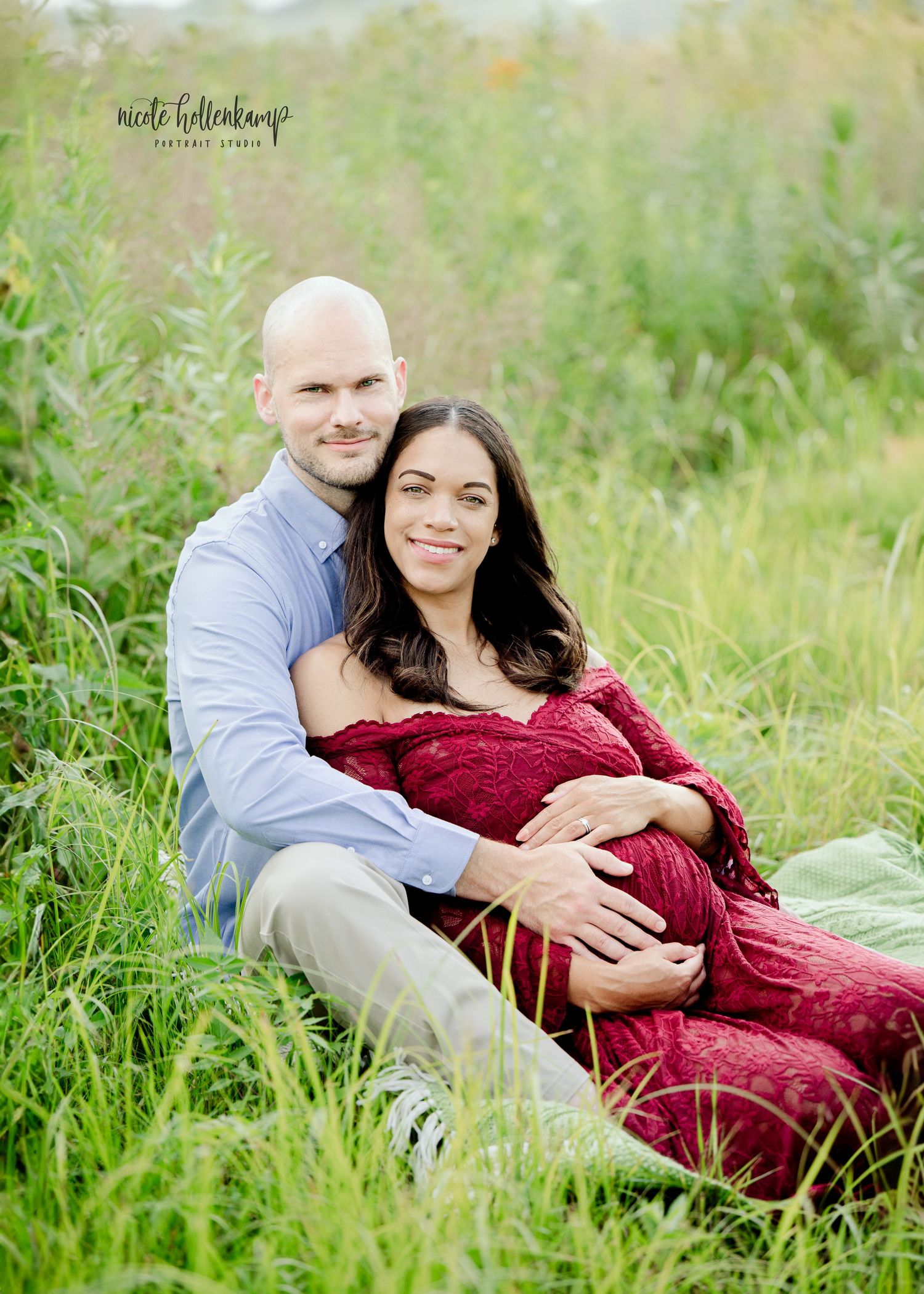 A couple sits in a lush green field, the woman in a red lace maternity gown cradling her baby bump.