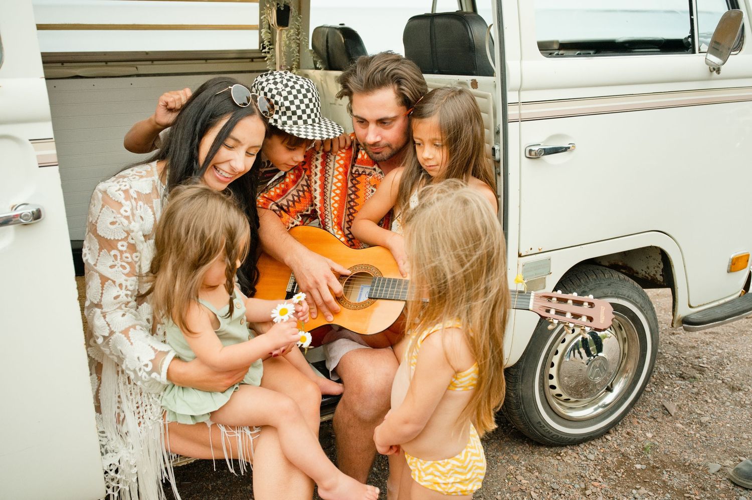 A bohemian family sits by a vintage van as a man plays acoustic guitar for two young girls and a woman.
