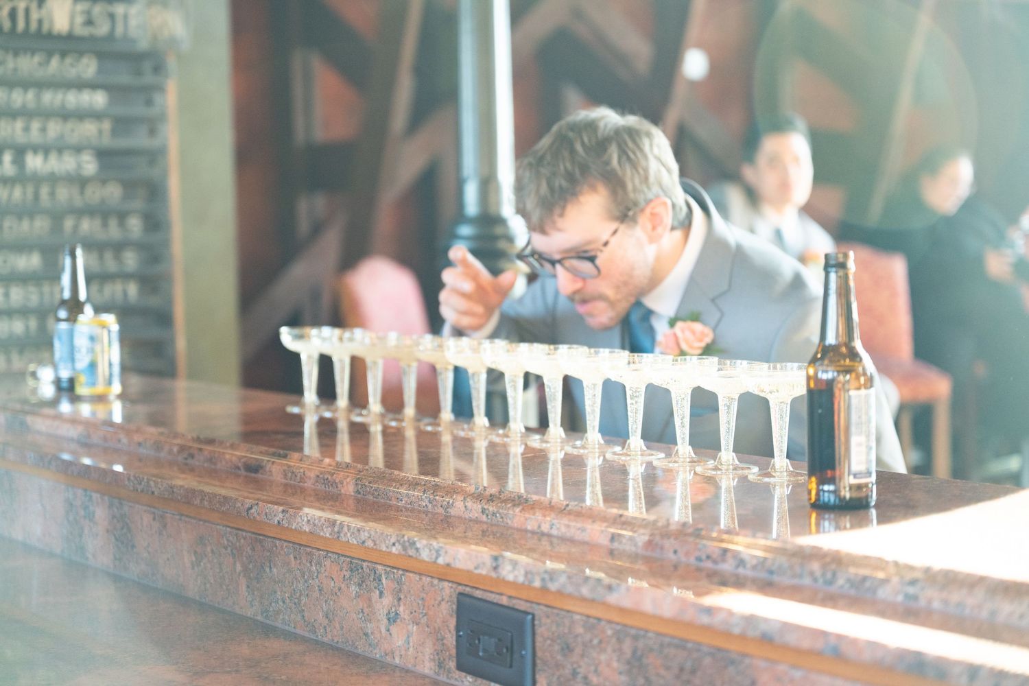 Man in suit carefully arranges row of shot glasses on polished wooden bar at wedding reception.