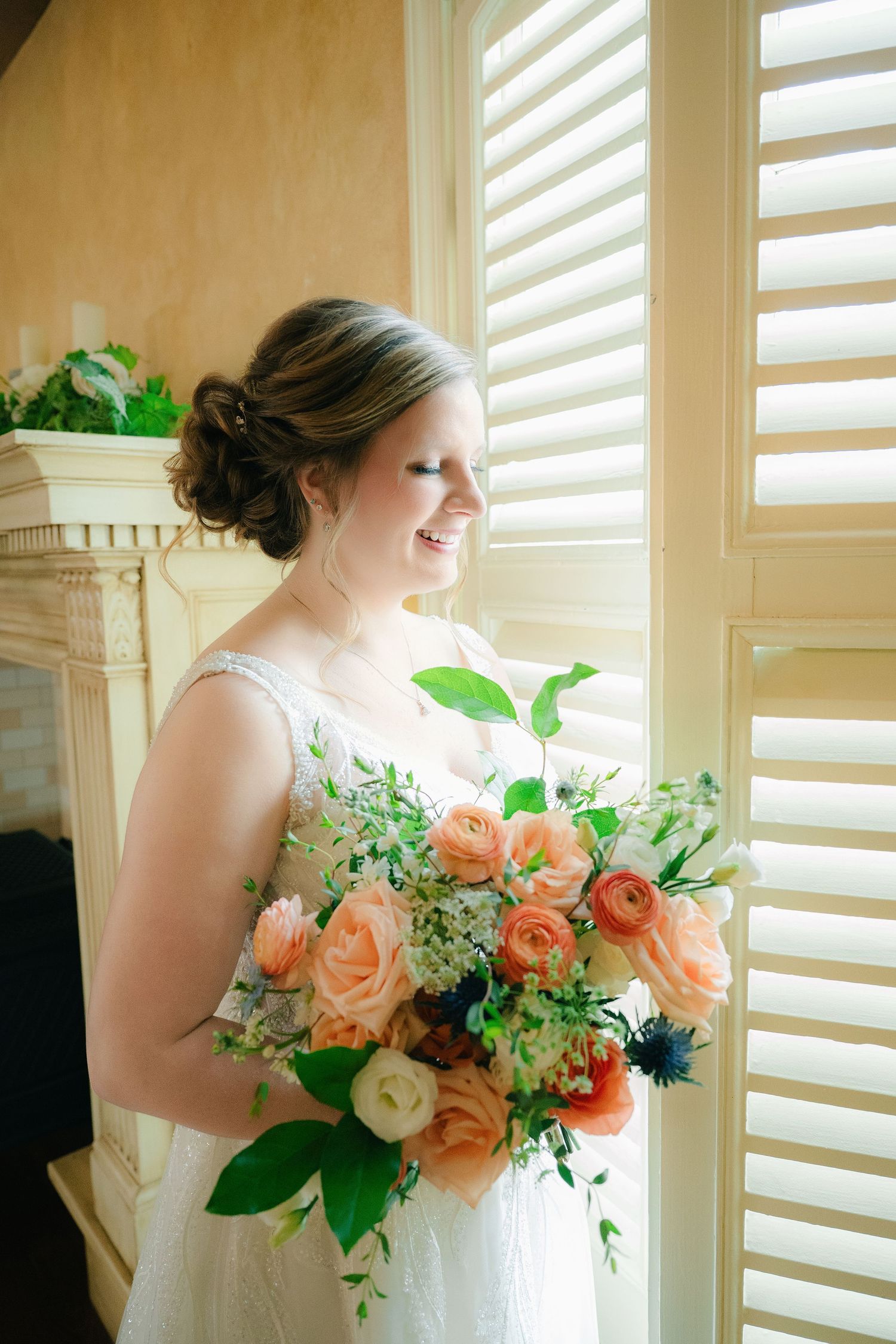 Smiling bride in a lace wedding dress holding a lush peach and white rose bouquet near white shuttered windows.