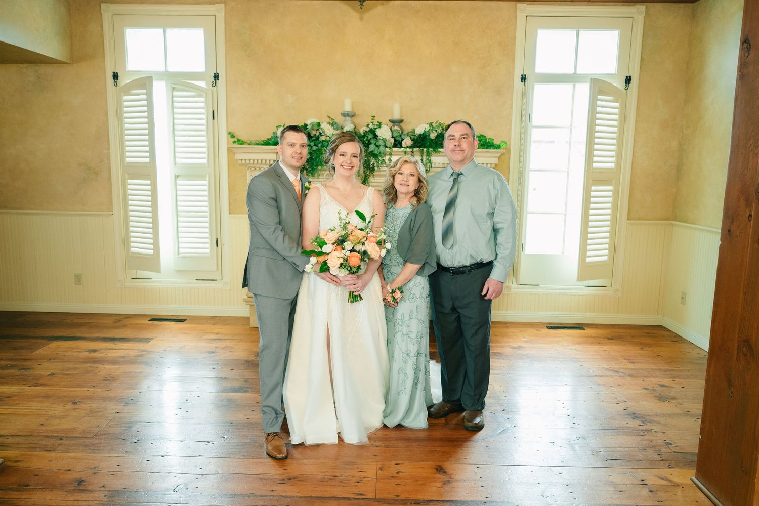 Bride with peach bouquet stands with three family members before floral fireplace backdrop.