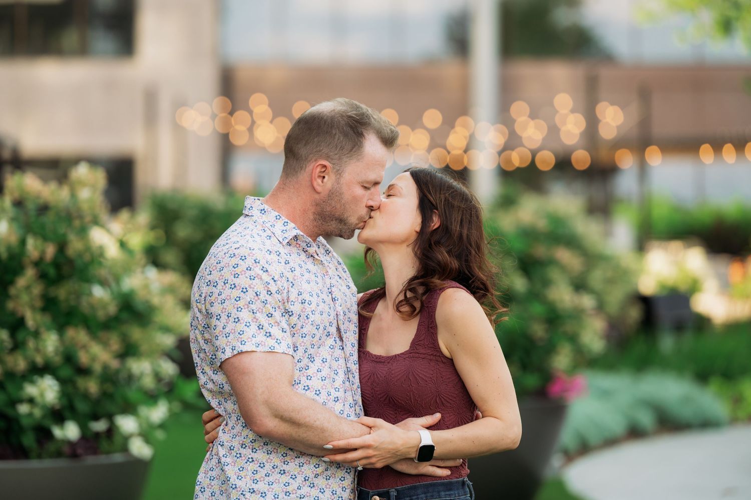 A couple shares a romantic kiss in a garden setting with soft lighting and blooming flowers in the background.