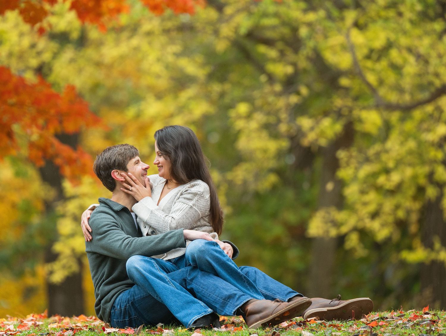 Two people laying together on fallen autumn leaves in a park with colorful trees in the background at Lake Como. Engagement Sessions MN.