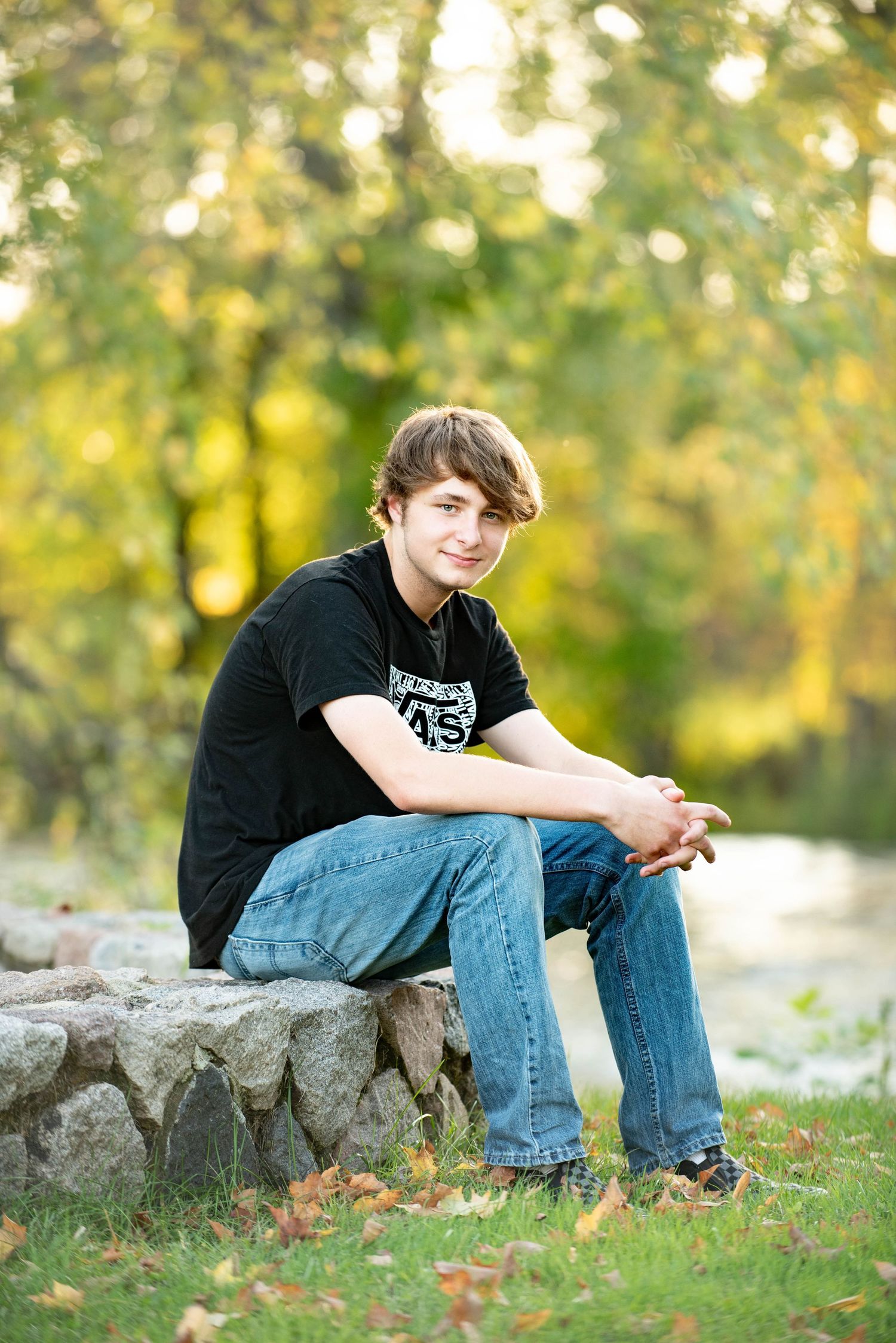 A person in a black shirt and jeans sits casually on a stone wall surrounded by autumn foliage in a park setting.