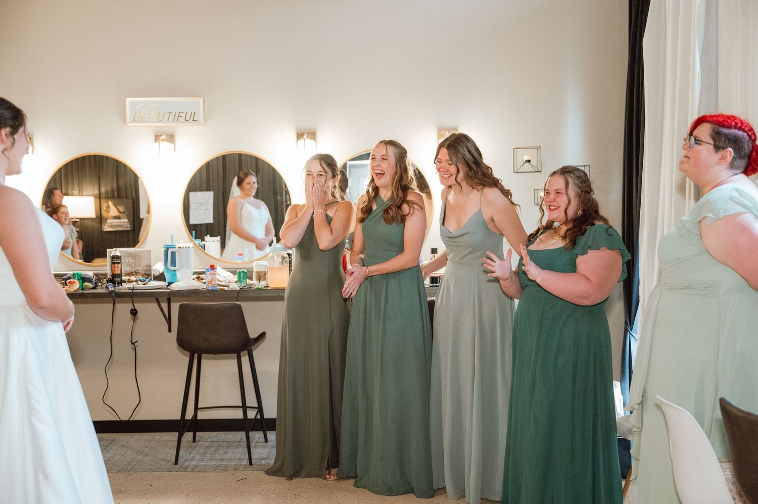 Wedding party members in sage green dresses react with surprise and joy during an emotional reveal moment.