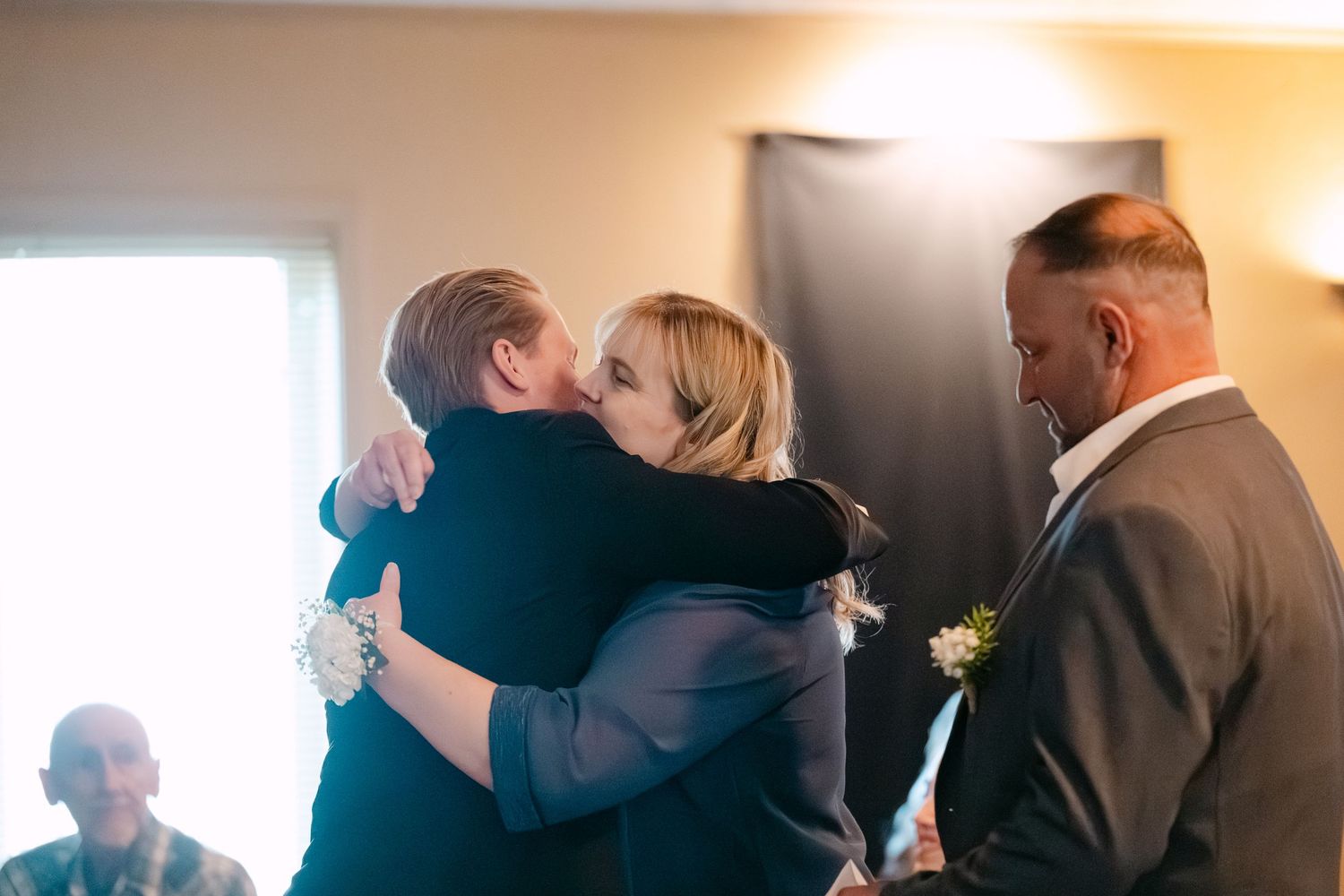 Two people share an emotional embrace during a wedding ceremony while others look on in a softly lit indoor setting.