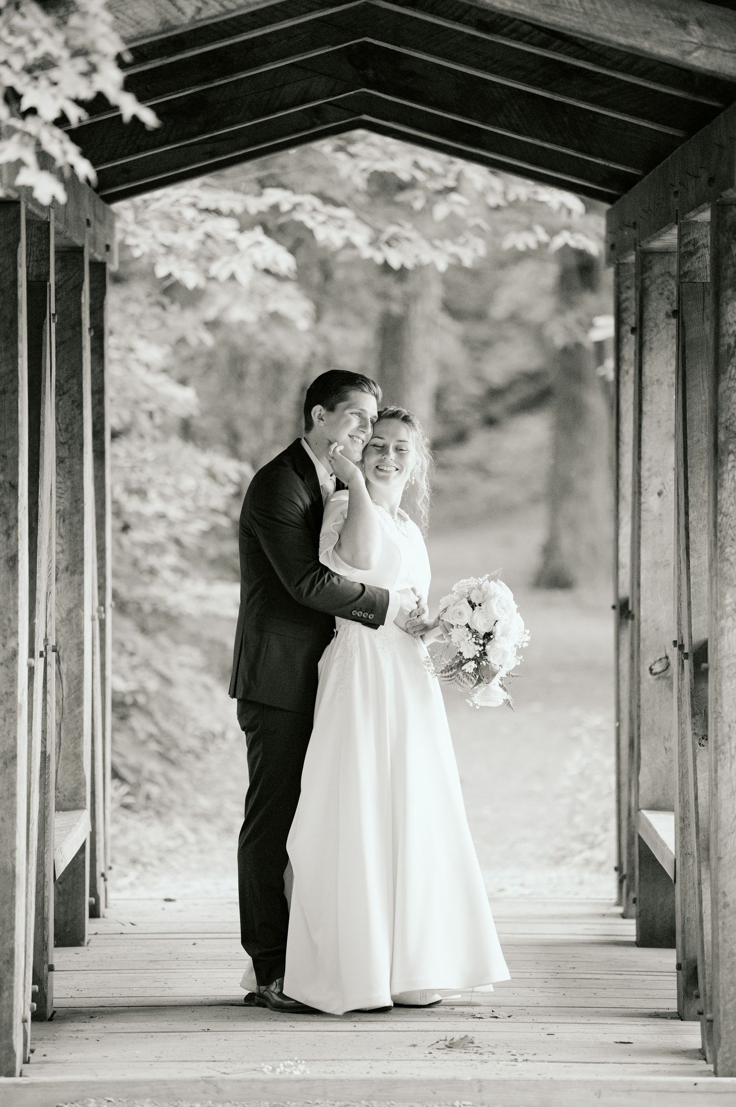 A romantic black and white wedding portrait captured under a covered wooden bridge walkway.