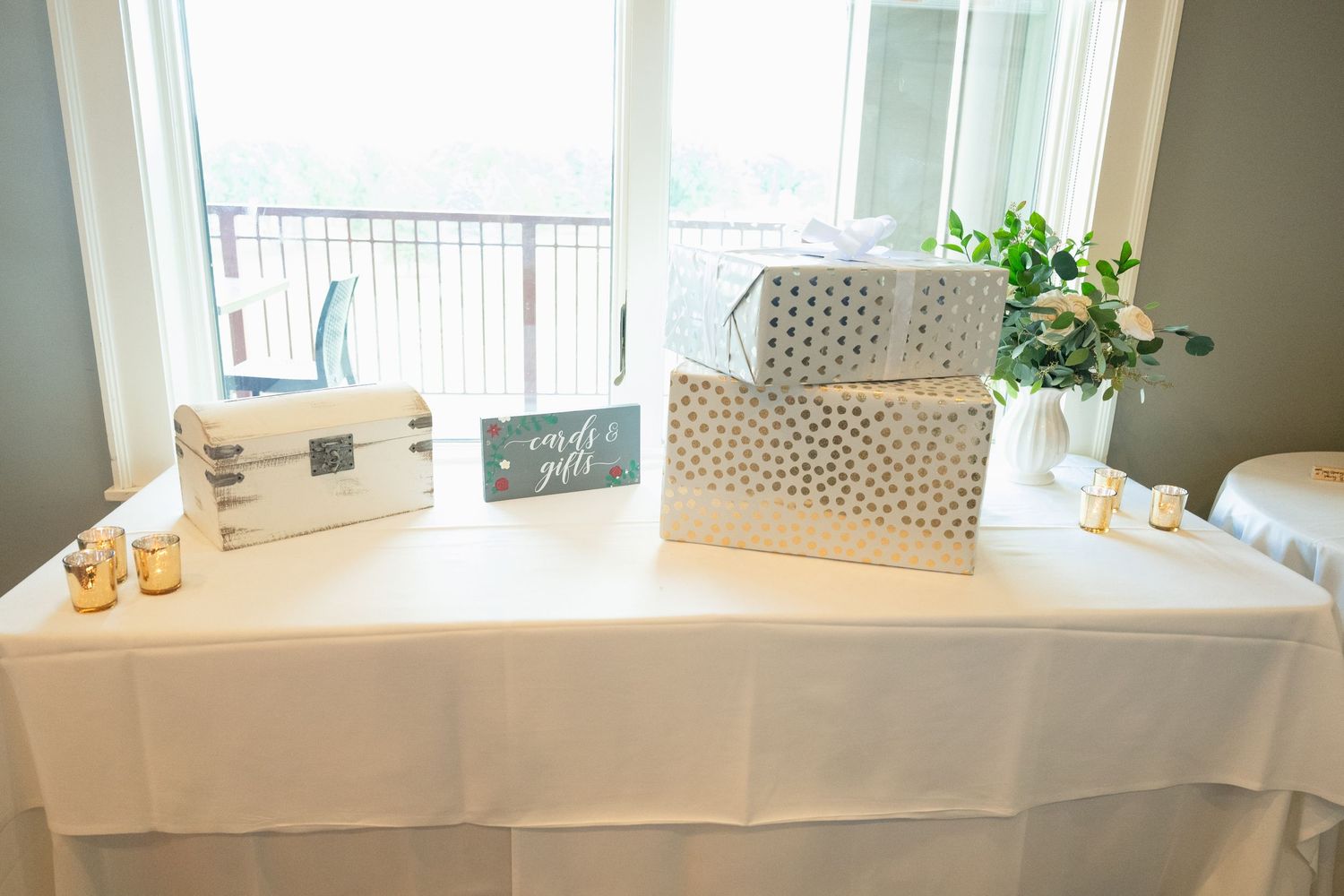 Wedding gift table with white decorative boxes, a card box, candles, and floral arrangement by a window.
