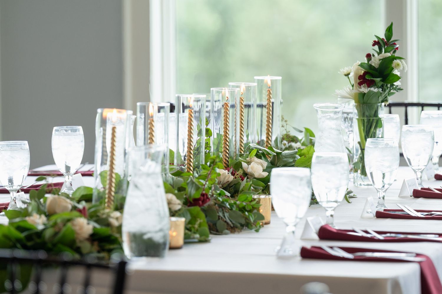Elegant wedding reception table with tall candles, greenery, white flowers, and burgundy napkins.