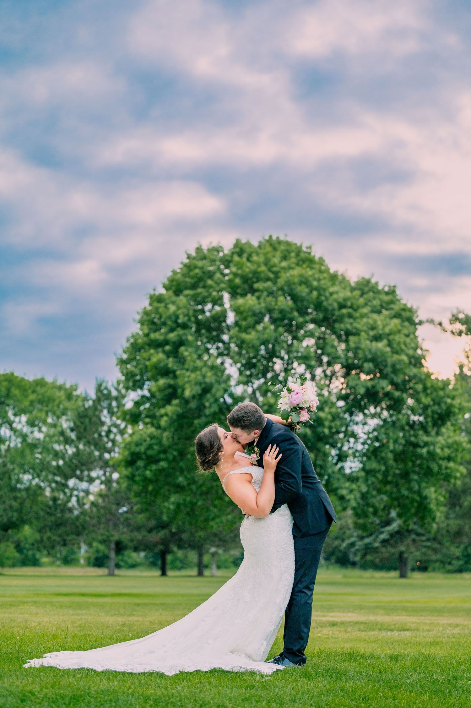 A bride and groom share a romantic dip kiss on a lush green lawn with a large tree and pastel sunset sky.