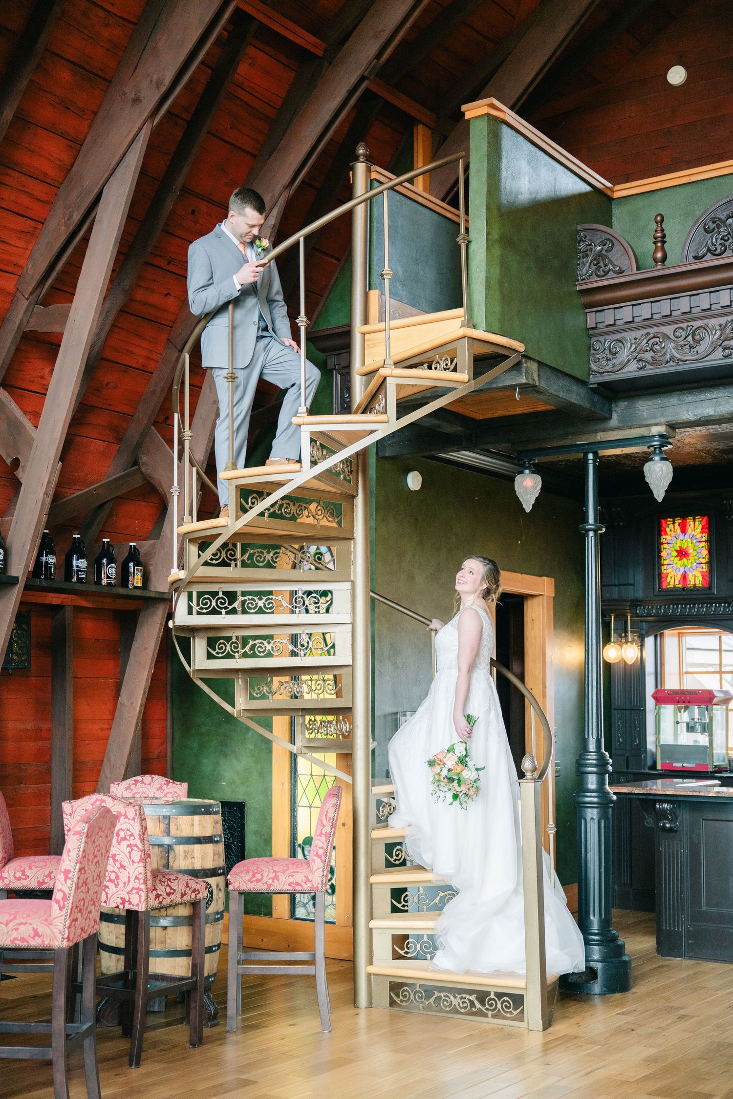 Bride and groom posing on ornate spiral staircase in rustic wedding venue with wooden beams and green walls.