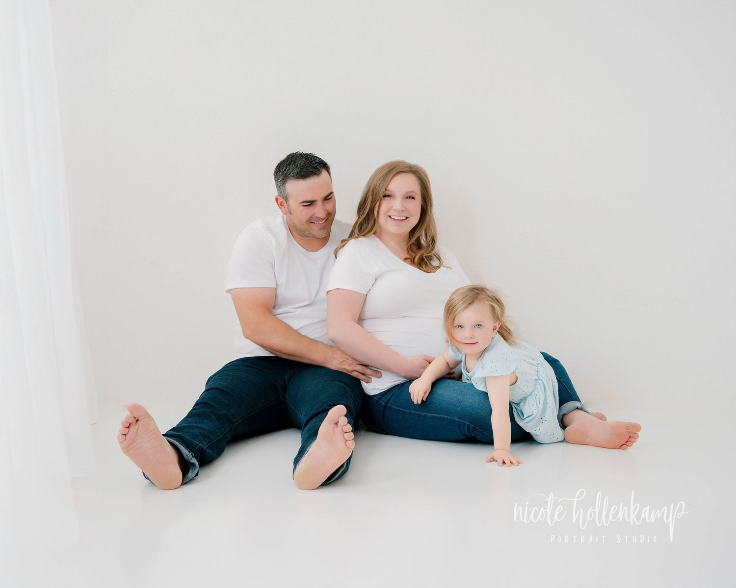 A family lounges together on a white bed wearing casual clothing in a bright minimalist setting.