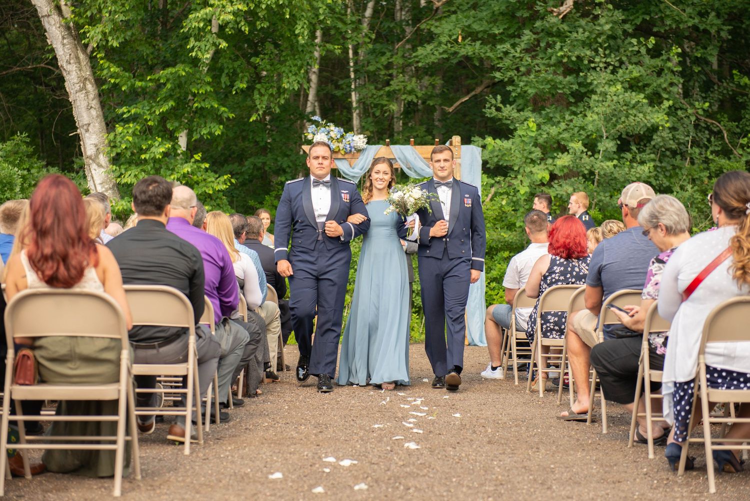 Wedding party walks down the aisle between folding chairs as guests look on at an outdoor forest ceremony. Zimmerman MN Weddings